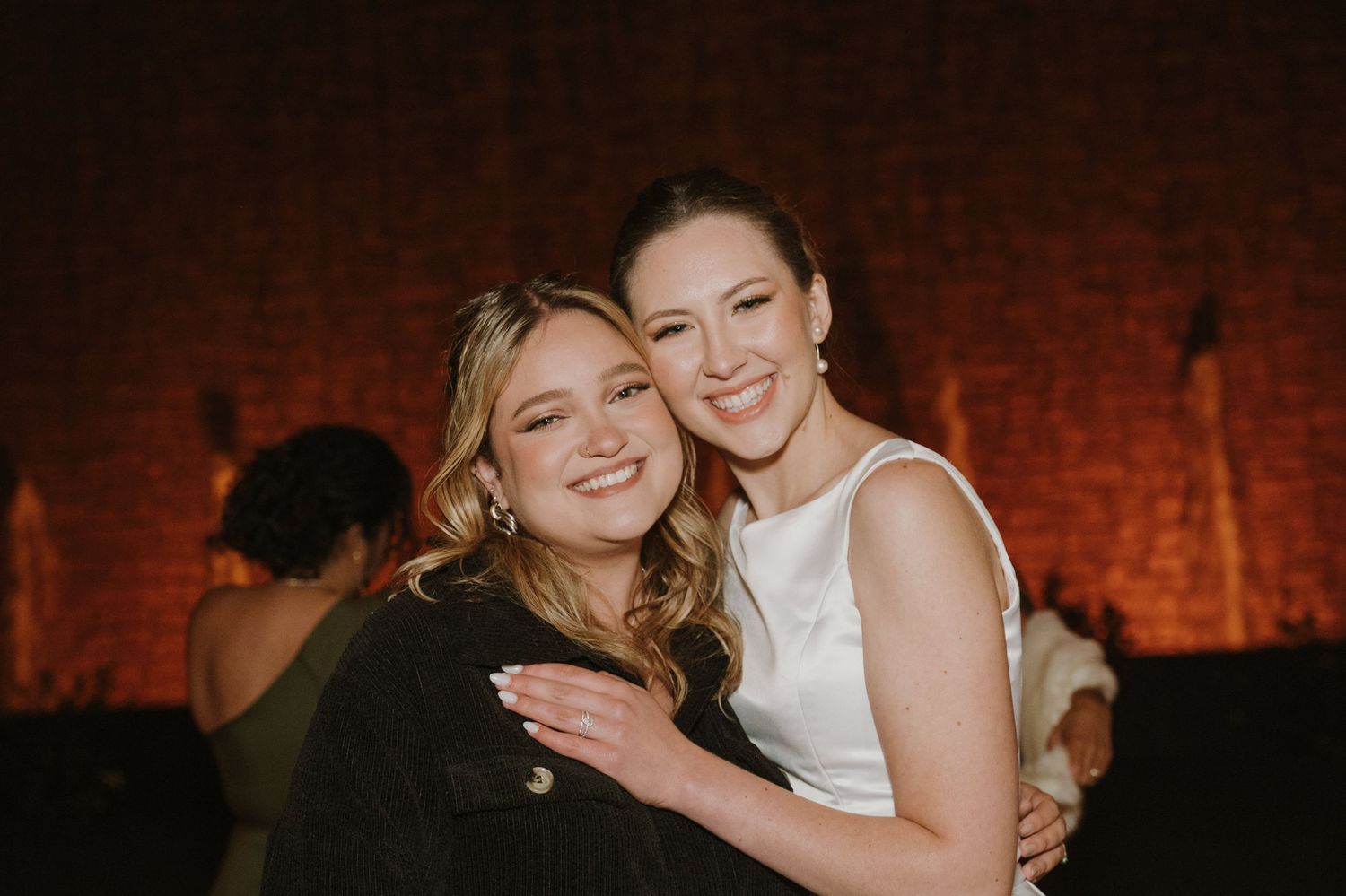 Two women smiling and embracing warmly at an evening wedding reception against a warm brick wall backdrop.