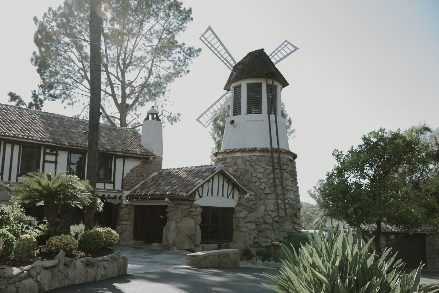 Stone building with a white windmill tower surrounded by lush greenery and trees in a scenic garden setting.