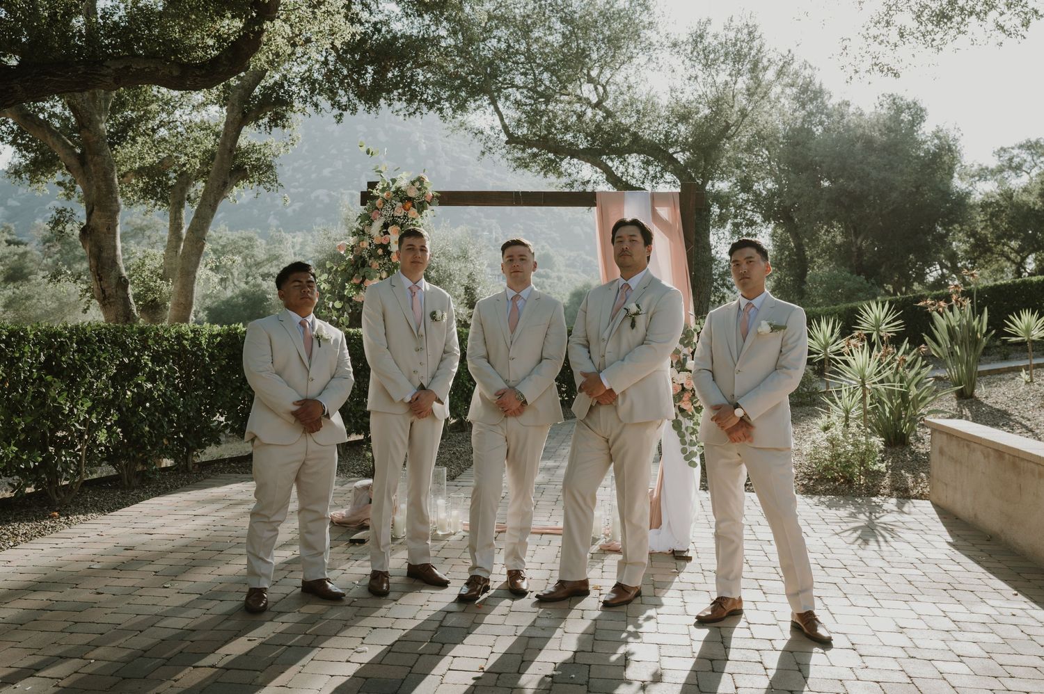 Groom and groomsmen pose in light suits before a floral arch in an outdoor garden.