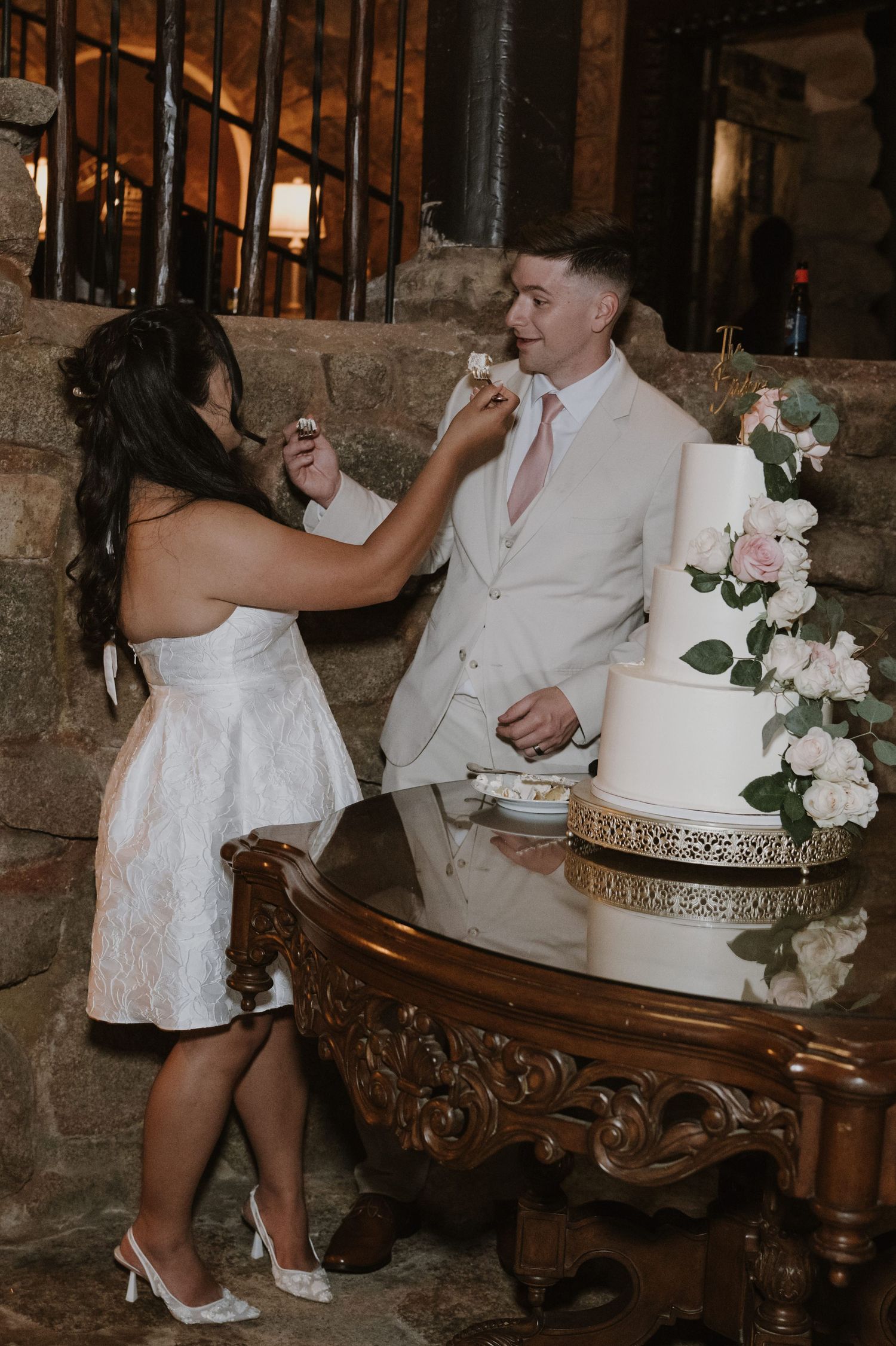 Bride feeding groom a piece of wedding cake beside a four-tier floral cake at their reception.
