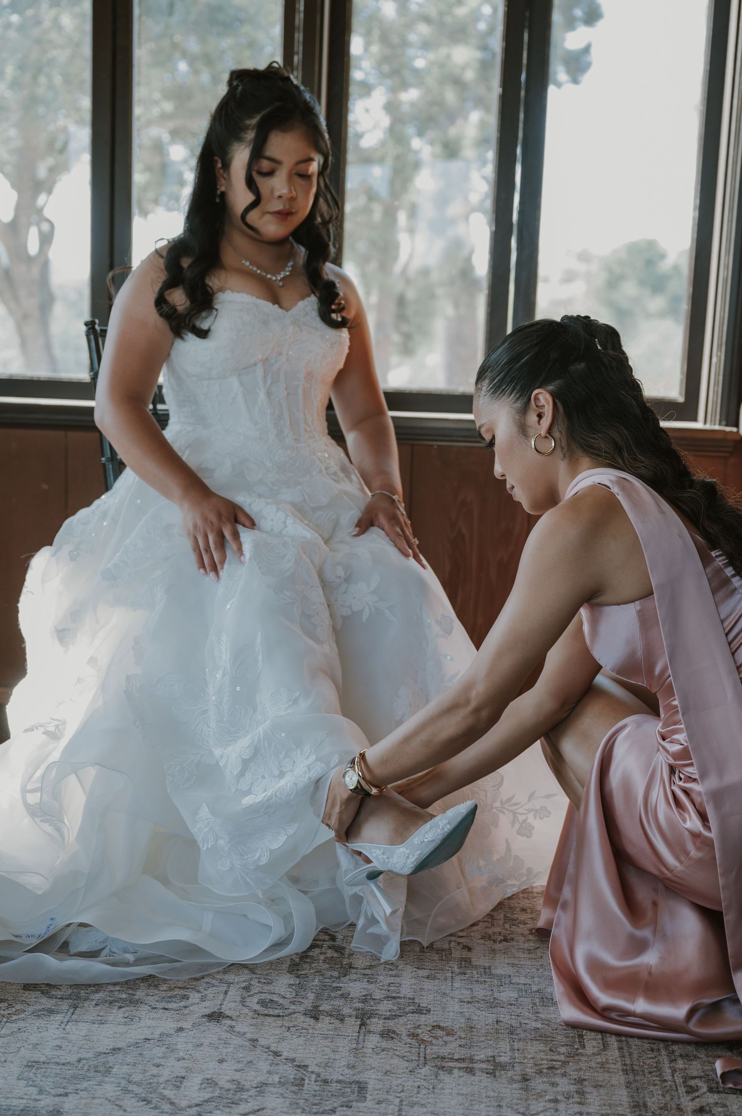 Bridesmaid in pink satin dress helps bride in white lace gown put on her wedding shoes.