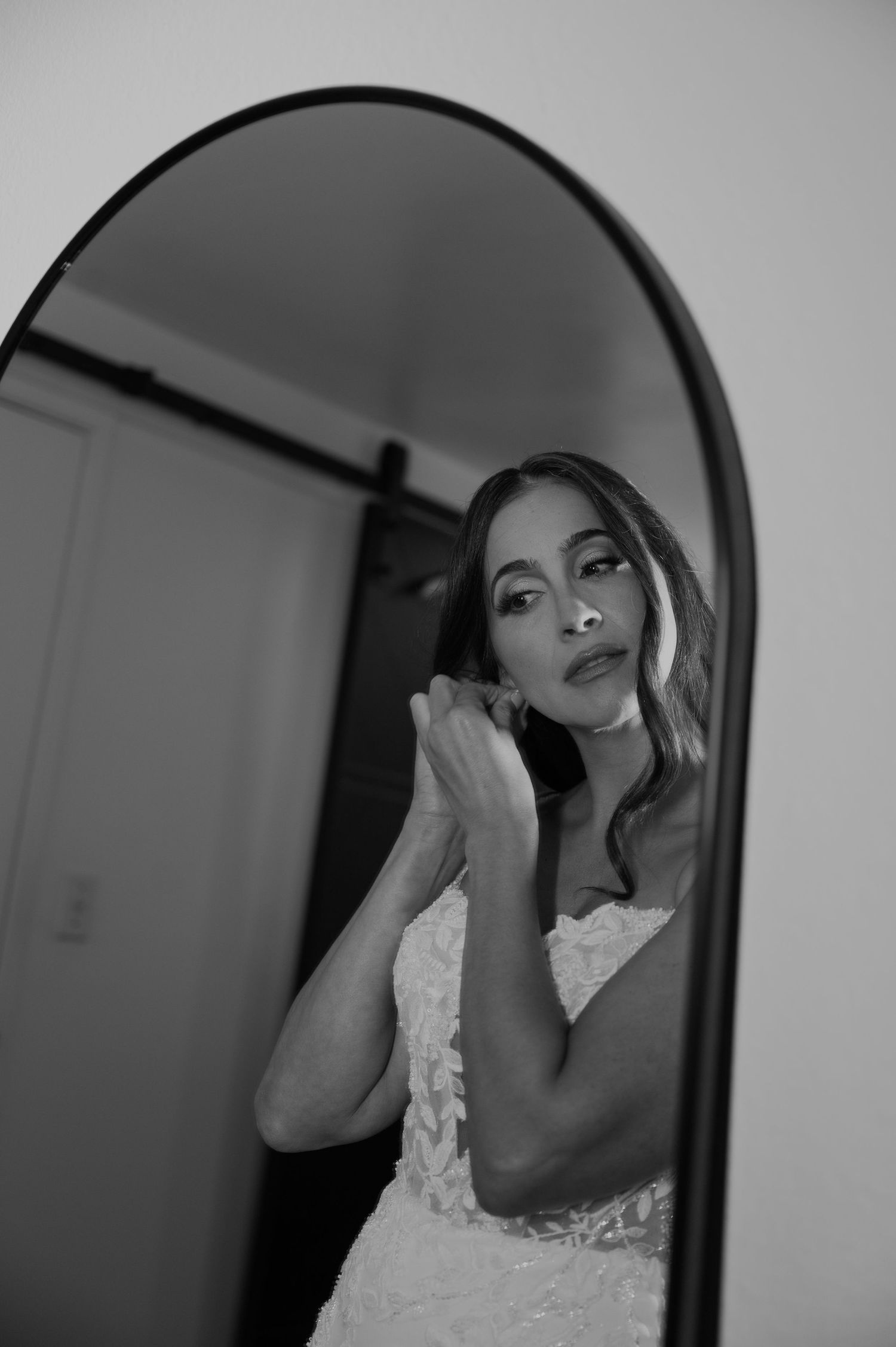 Black and white photo of a bride in a white dress adjusting her earring while looking in an arched mirror.