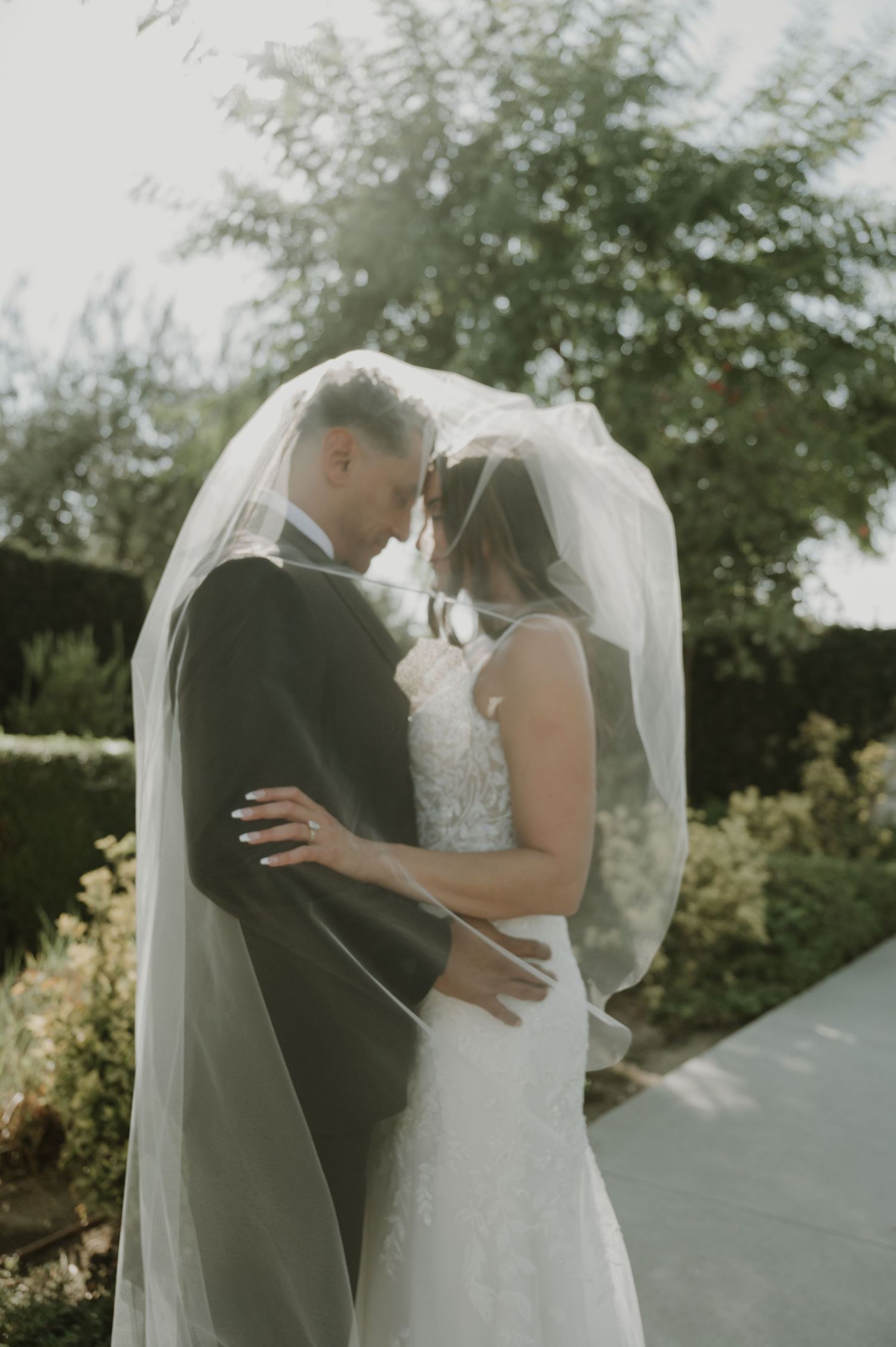 Bride and groom share an intimate moment under a flowing veil in a sunlit garden on their wedding day.