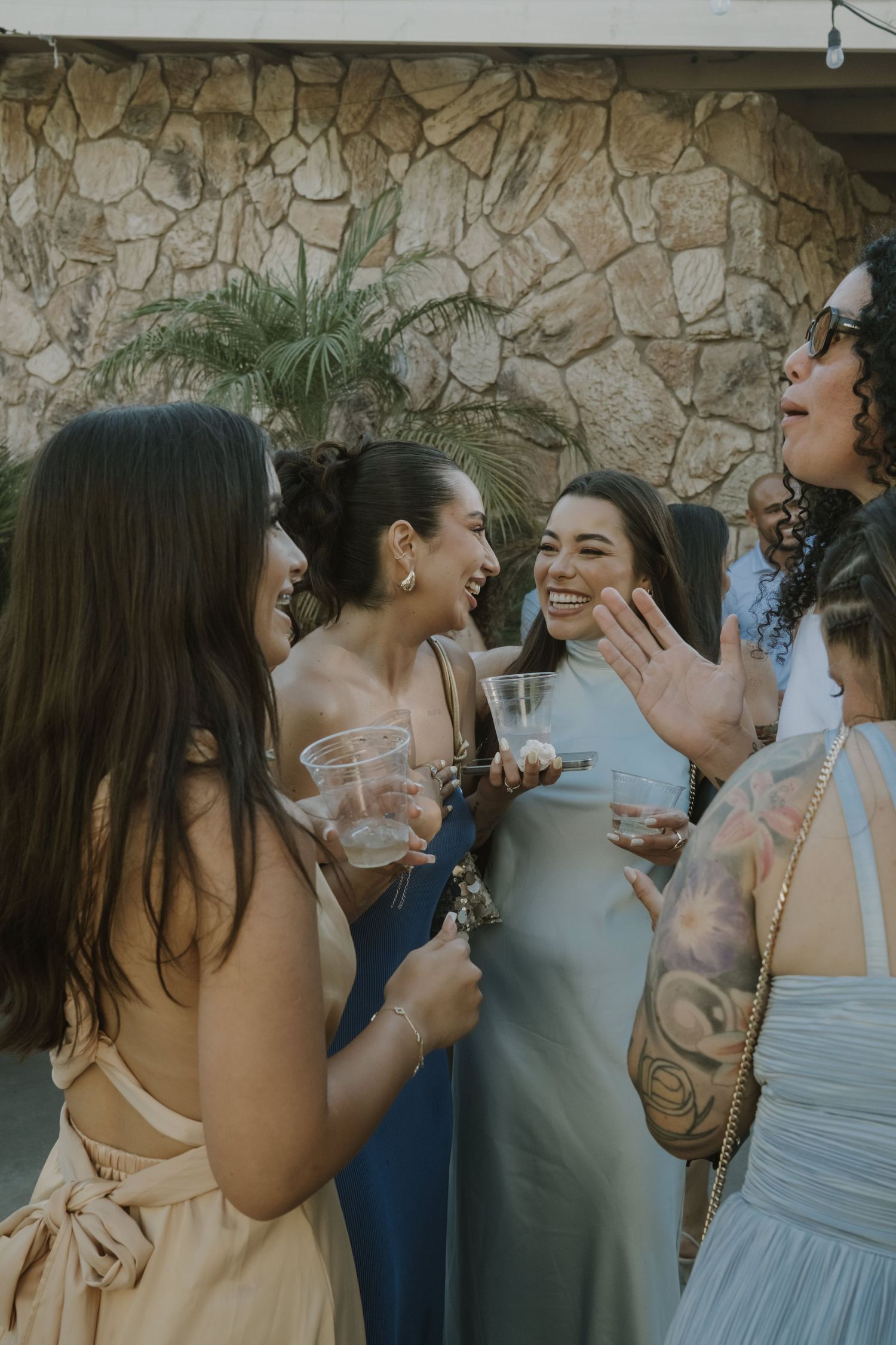 Group of young women laughing and holding drinks at an outdoor wedding celebration.