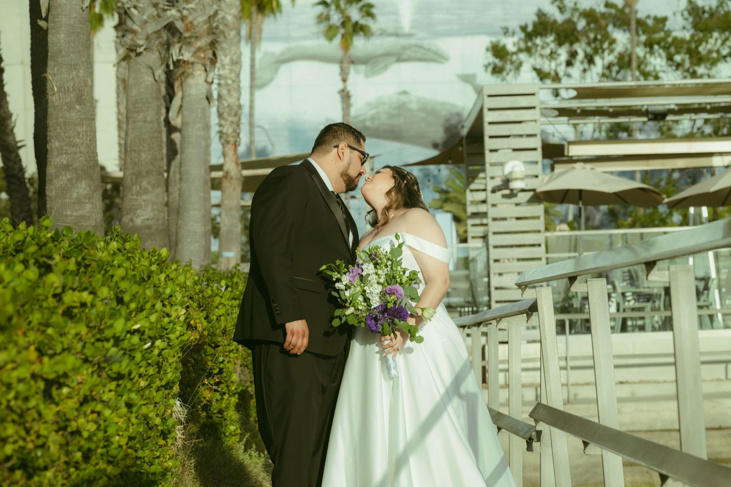 Bride and groom share a romantic moment outdoors, bride holding purple and white bouquet in elegant wedding gown.