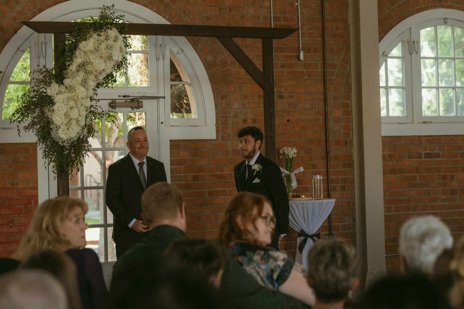 Groom and officiant stand at wedding altar with white floral arch in brick venue, guests seated in foreground.