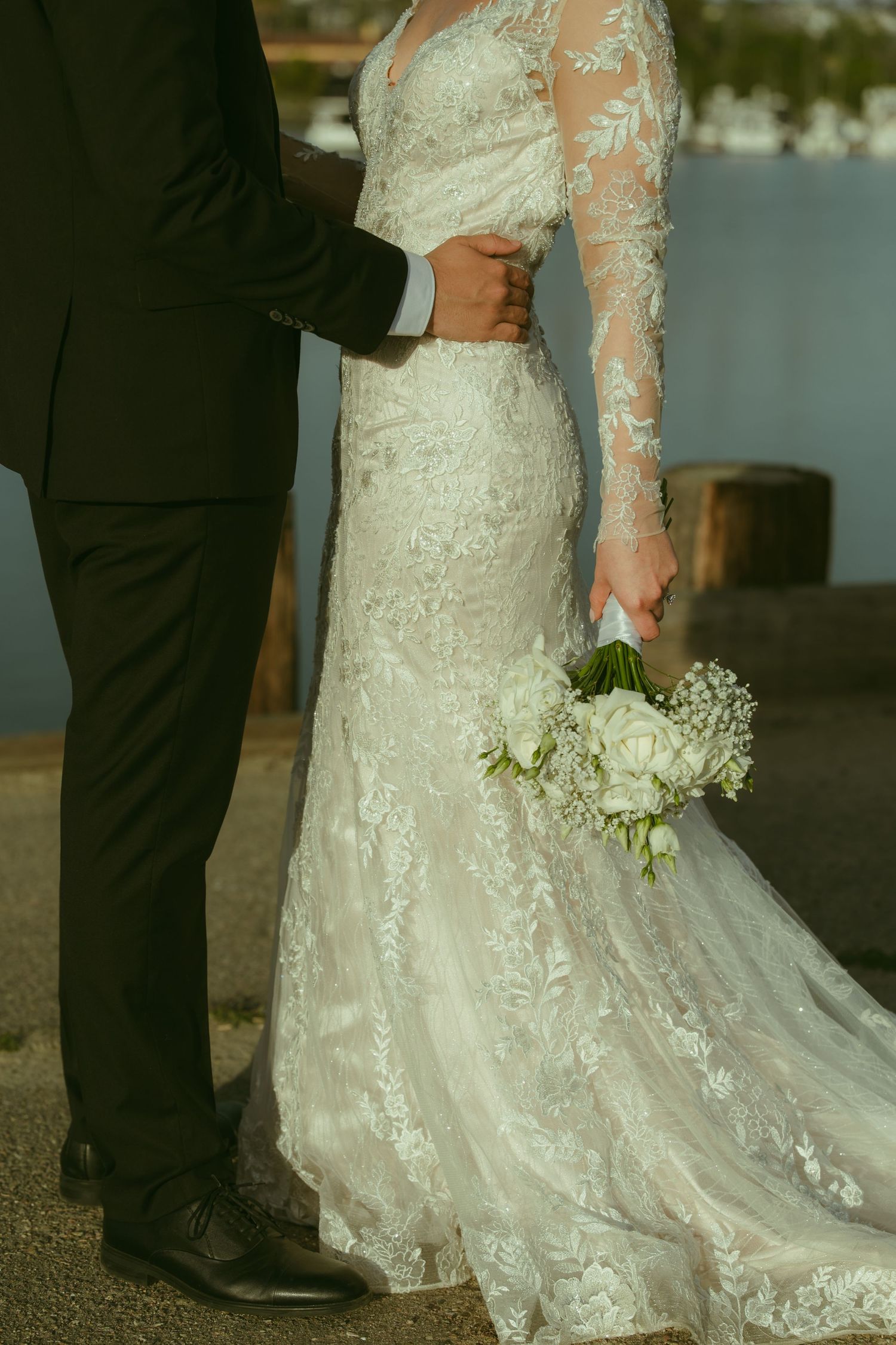 Close-up of bride in long-sleeve lace wedding gown holding white floral bouquet beside groom in black suit.