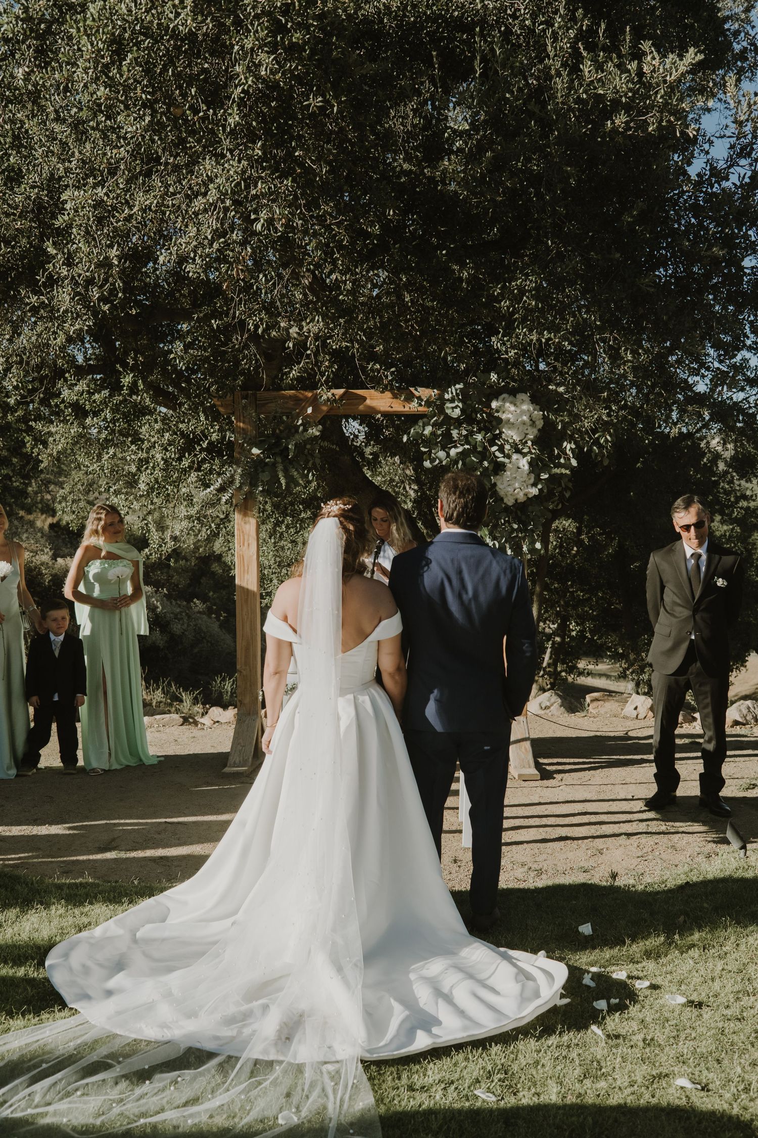 Bride in white gown with veil stands at outdoor wedding ceremony altar beneath large tree with bridesmaids in mint dresses.
