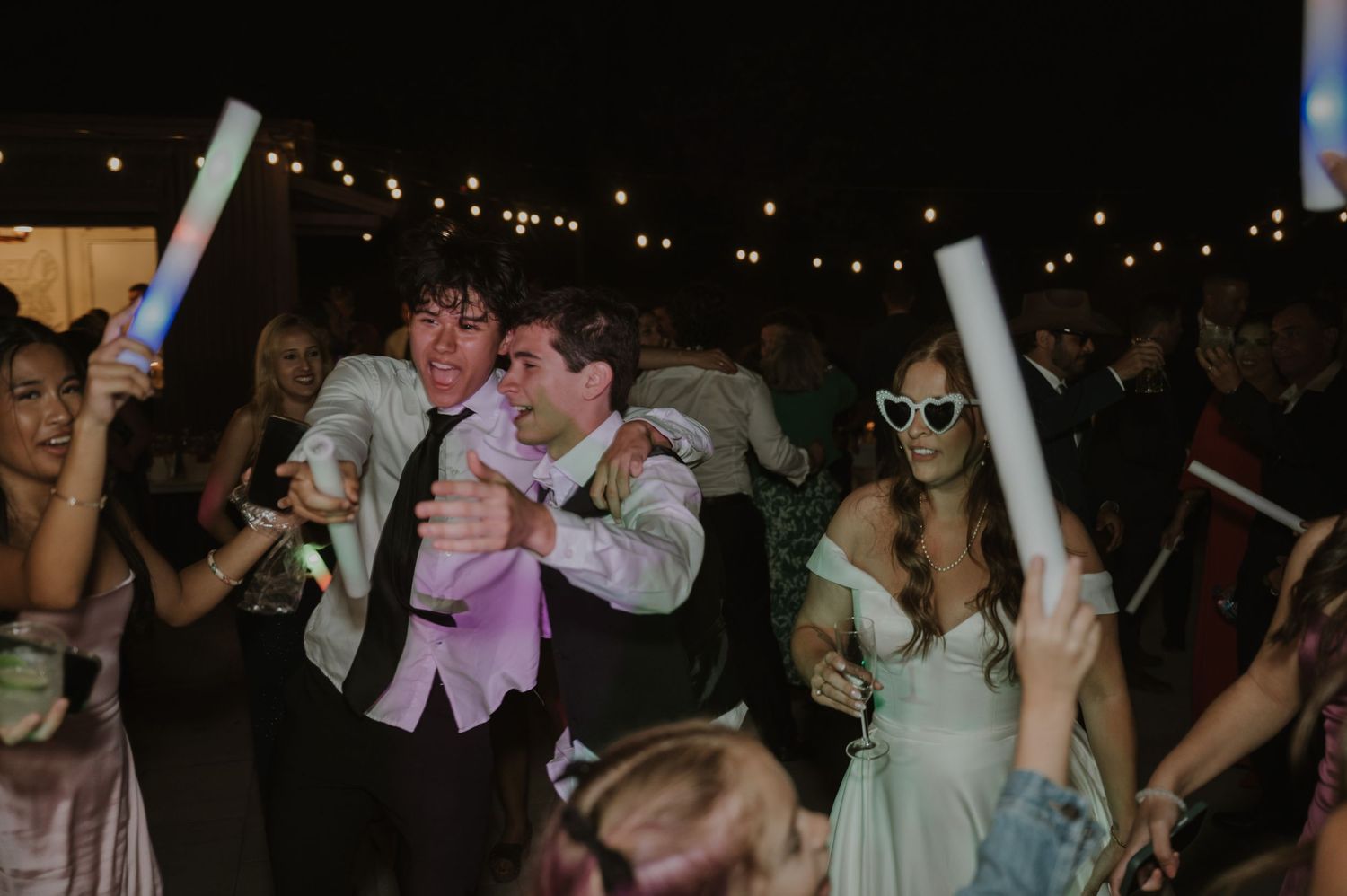 Excited wedding guests dancing with glow sticks at lively outdoor wedding reception party at night.