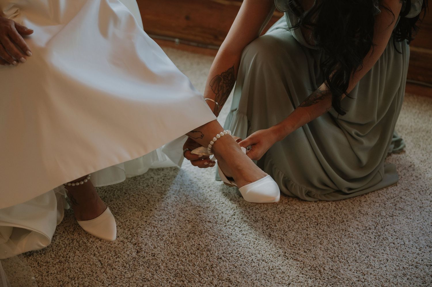 Bridesmaid in sage green dress kneeling to help bride put on white heels on carpeted floor.