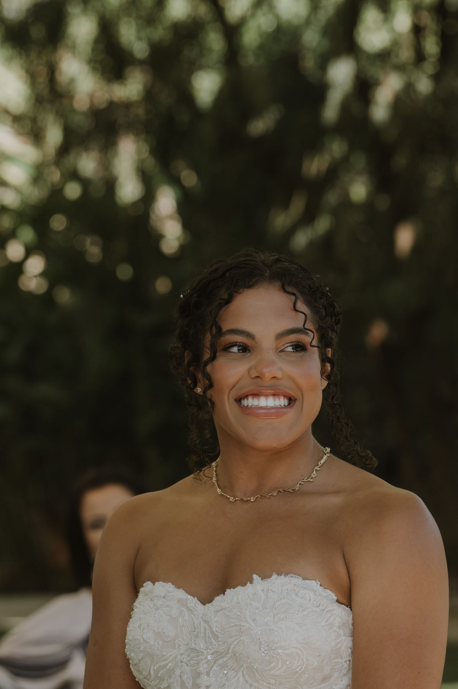 Smiling bride in strapless white lace gown with curly hair and gold necklace at outdoor wedding ceremony.
