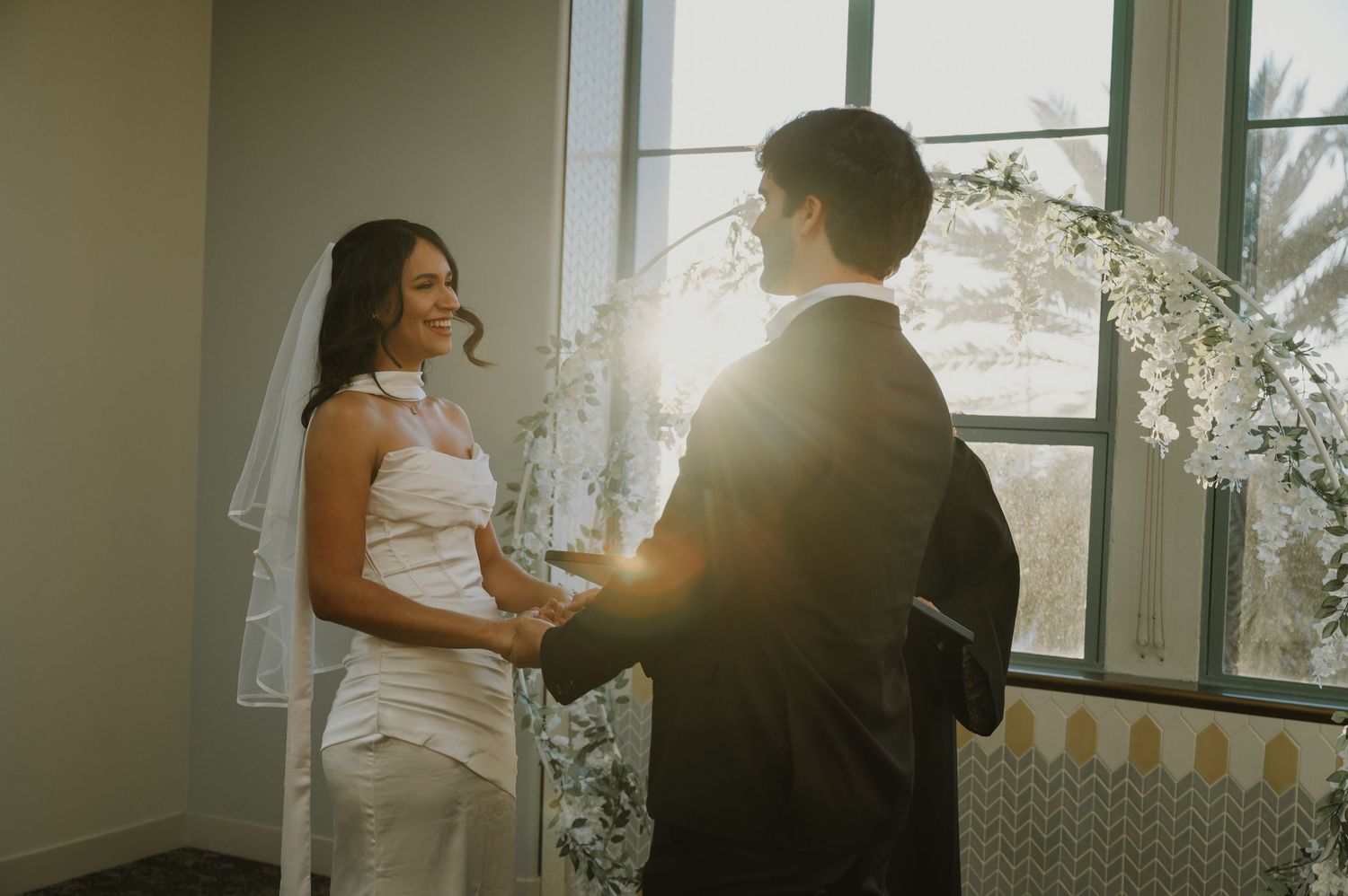Sunlit wedding photos show tender moments between newlyweds by large windows.