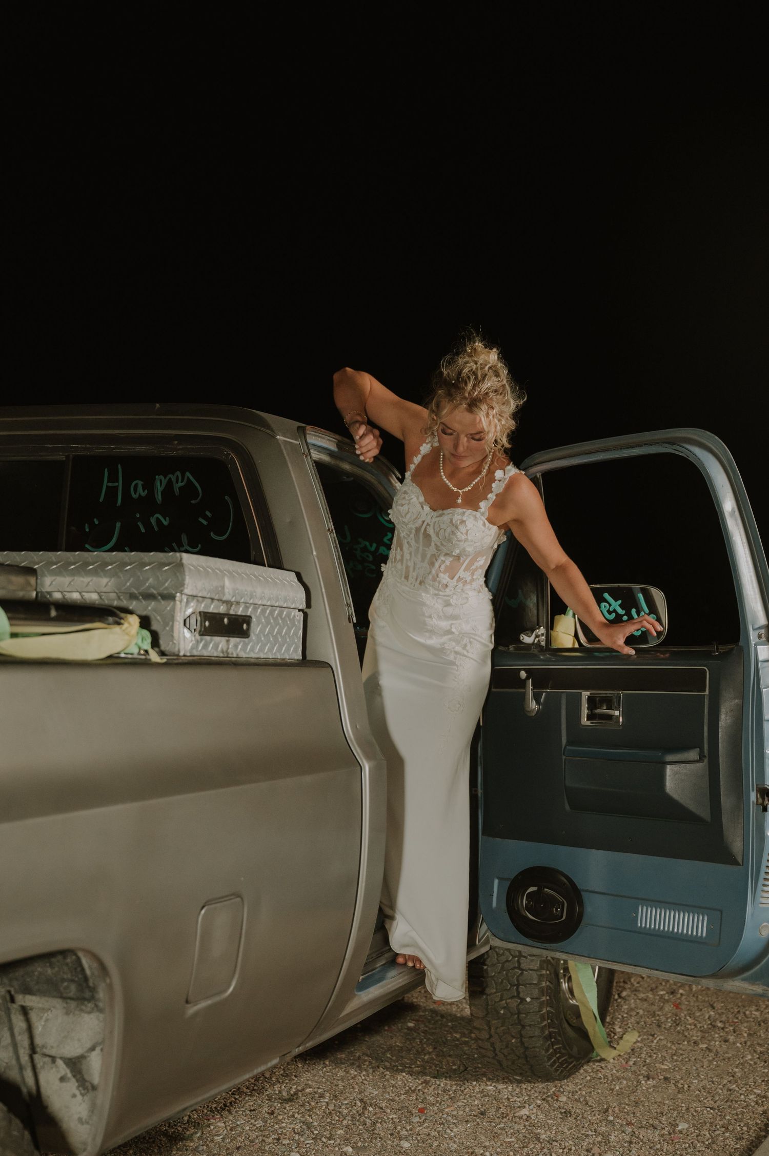 Person in white wedding dress enters a vintage car during evening celebrations.