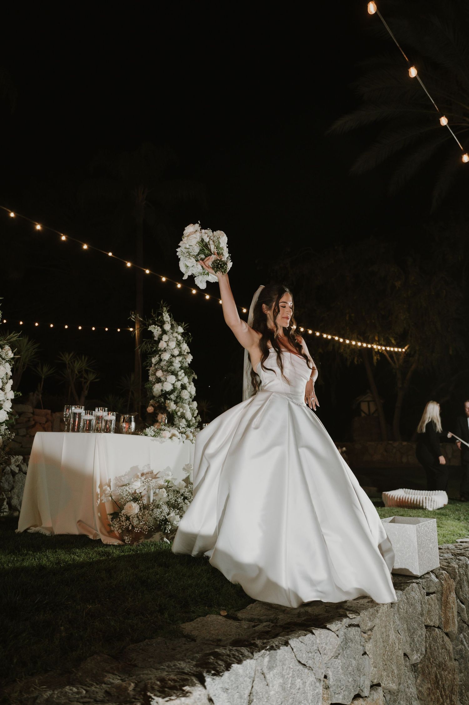 Outdoor wedding celebration with string lights and stone wall backdrop at night.