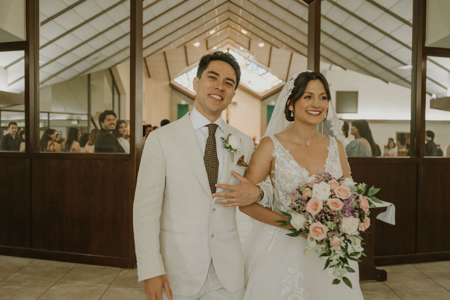 Smiling bride and groom pose together in a chapel, she holds a pink bouquet, he wears a white suit with brown tie.