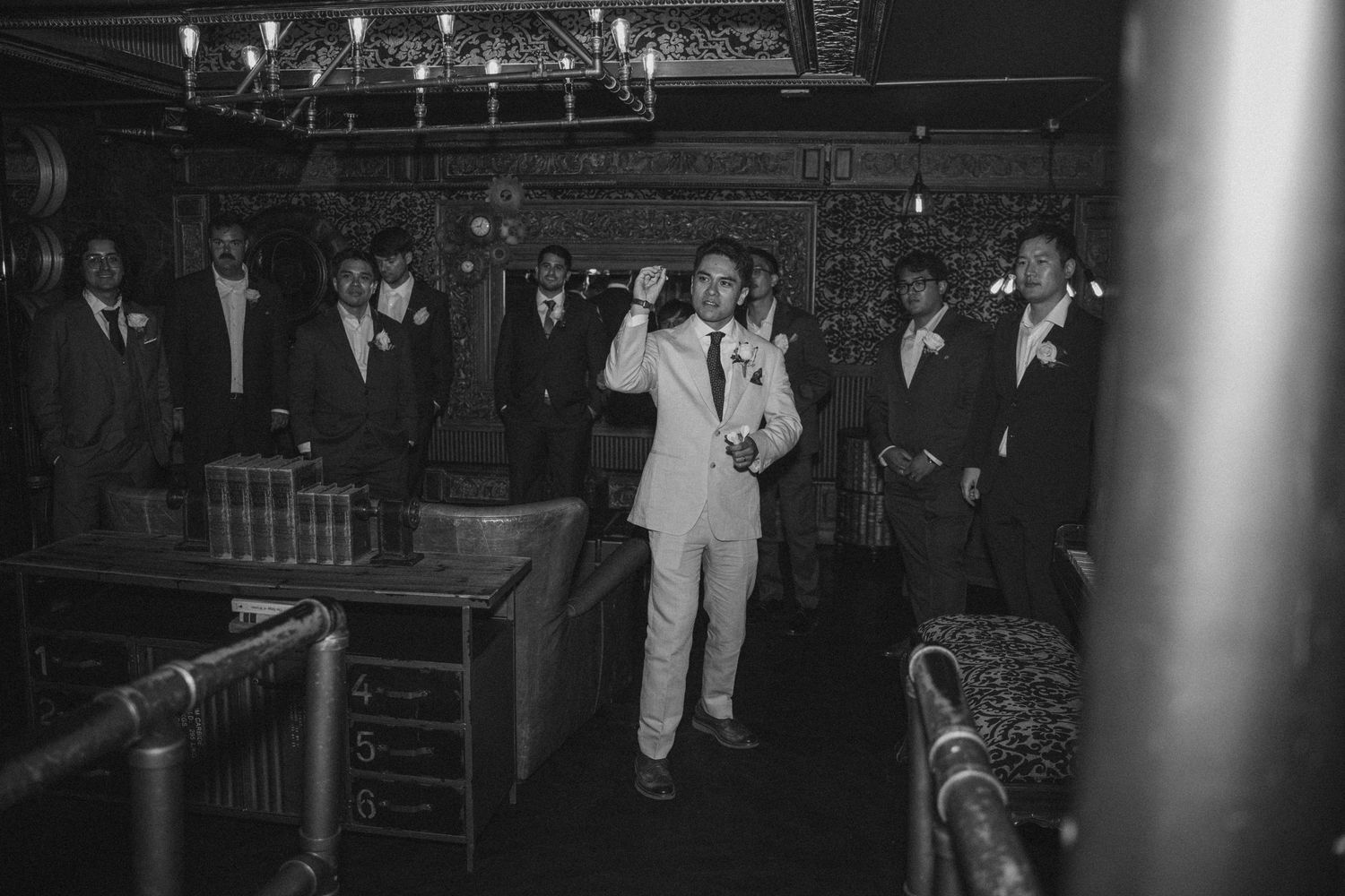 Groom in light suit addresses groomsmen in elegant dark wood room, black and white photo.