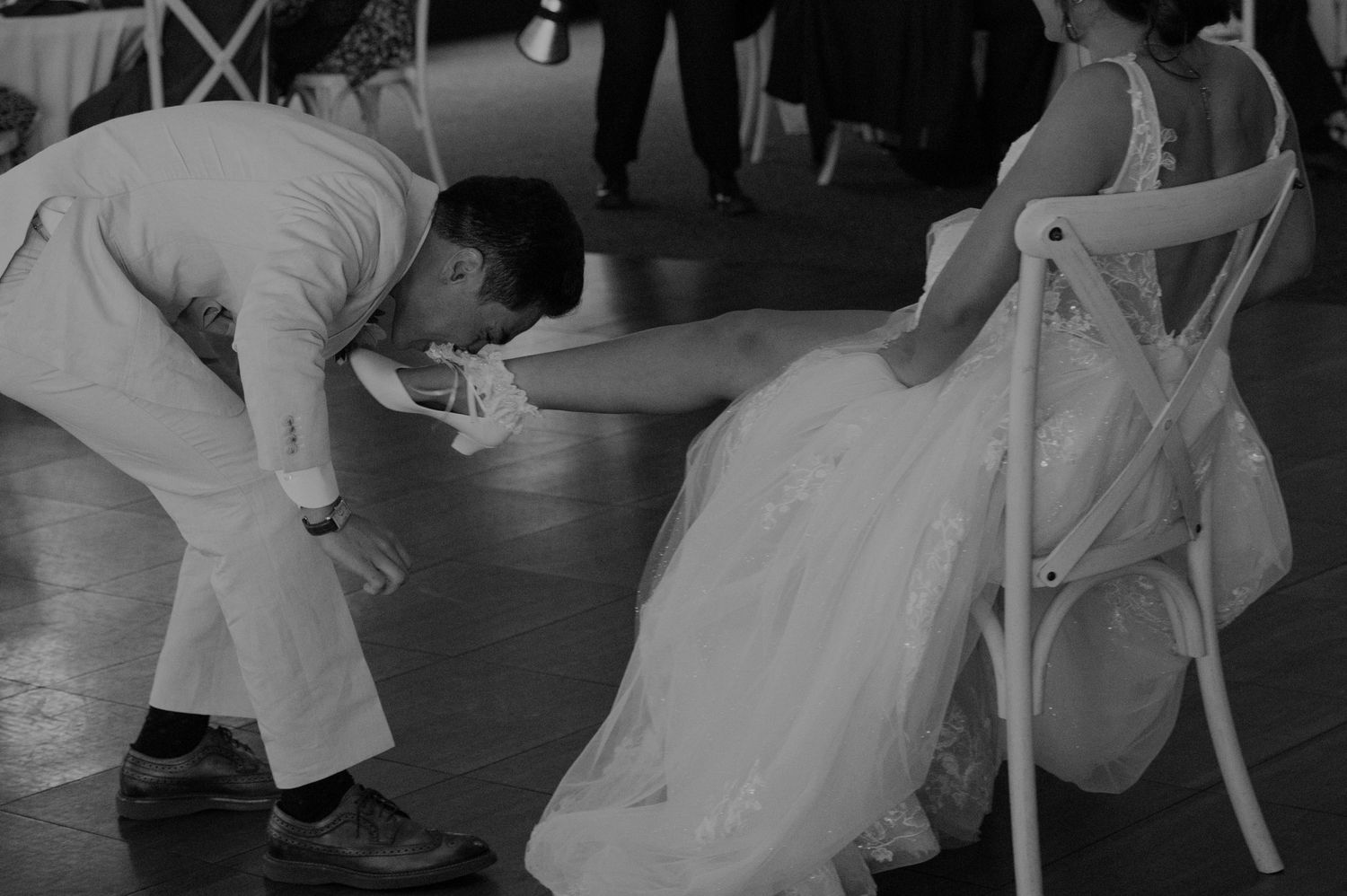 Black and white photo of groom retrieving the bridal garter from beneath the bride's wedding dress.