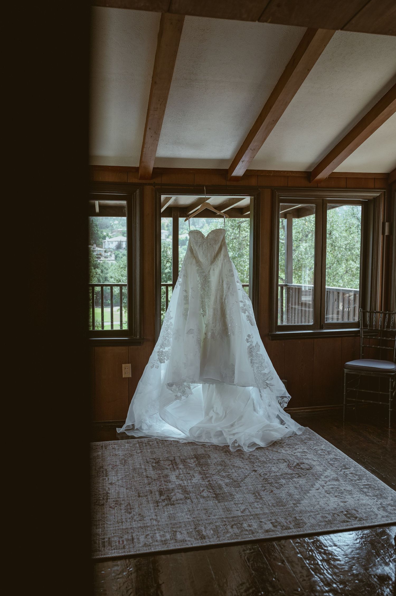 Elegant white wedding gown with lace details hanging in a rustic wood-beamed room with large windows.