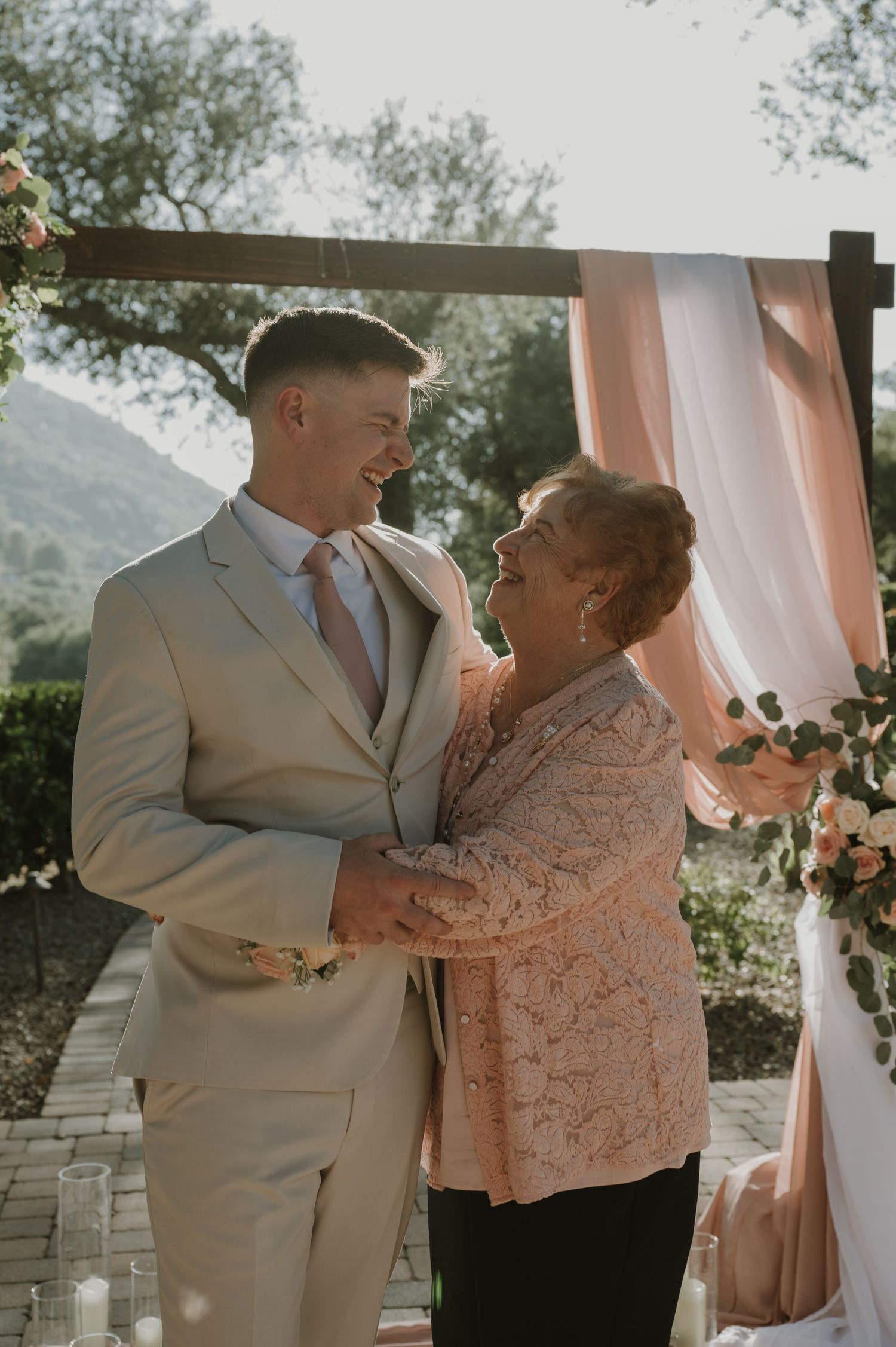 Groom in tan suit embracing older woman at outdoor wedding ceremony beneath floral arch.