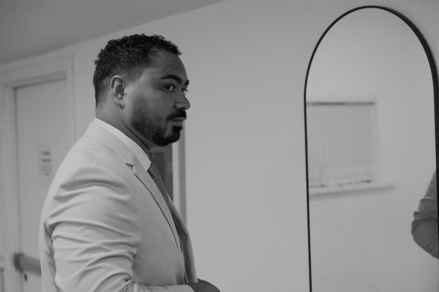 Black and white portrait of a man in a white blazer standing thoughtfully in a modern interior setting.