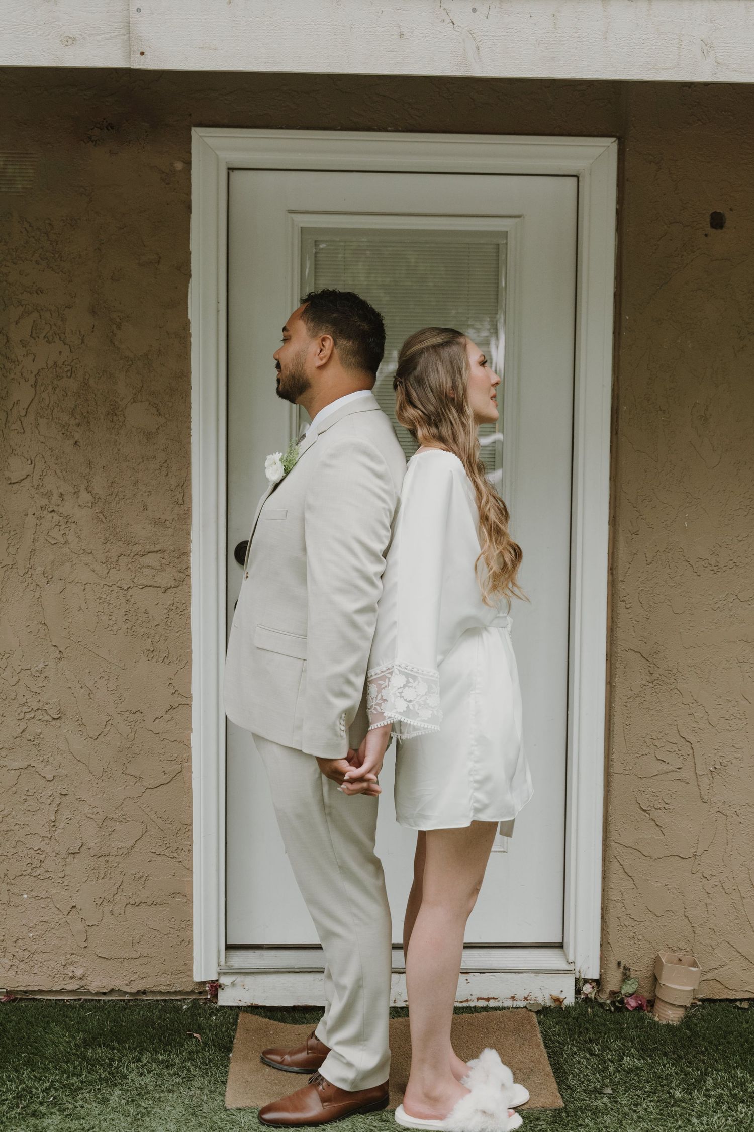 Bride and groom standing back-to-back in white and beige wedding attire at a white door.