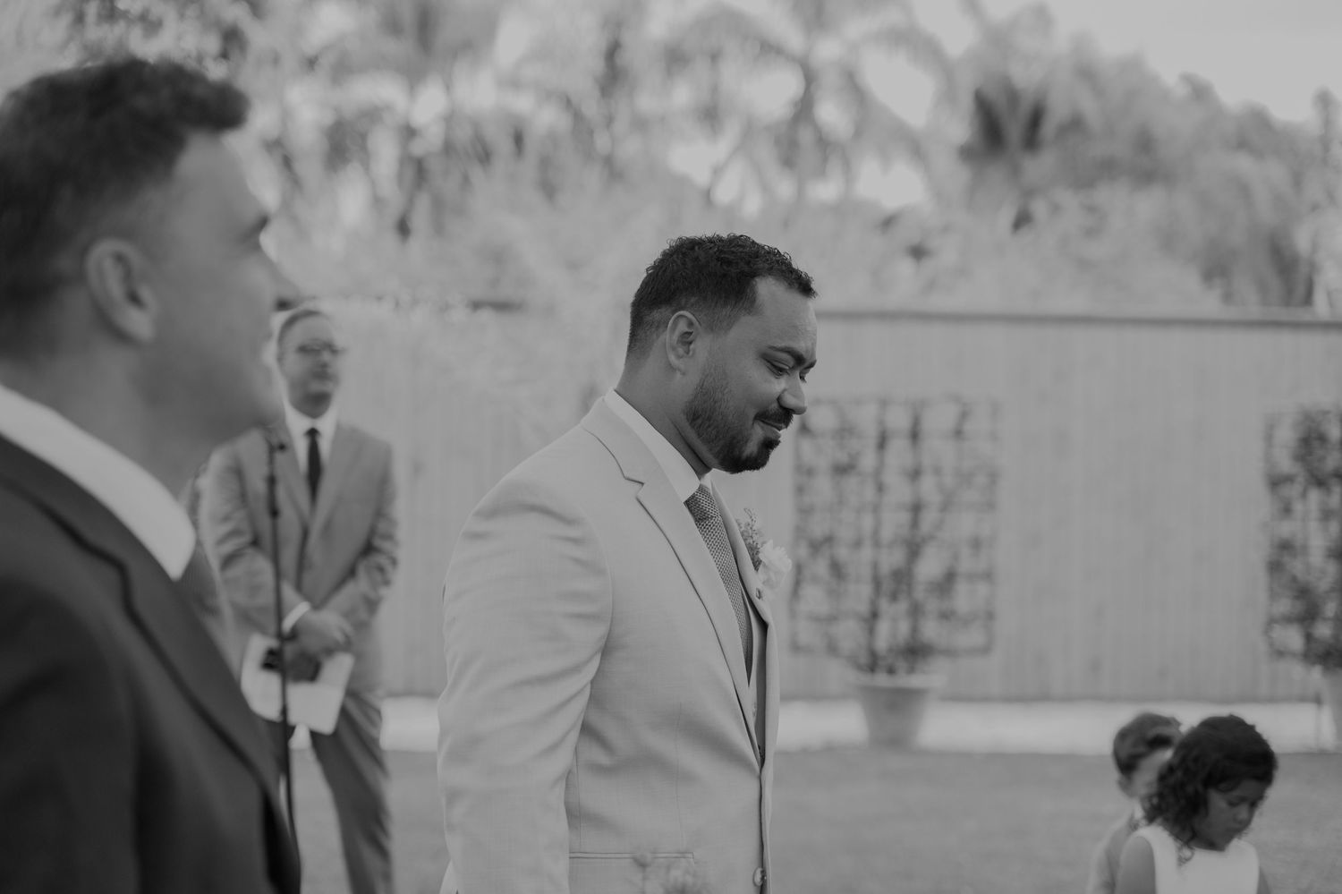Black and white photo of a groom in a light suit smiling at an outdoor wedding ceremony.