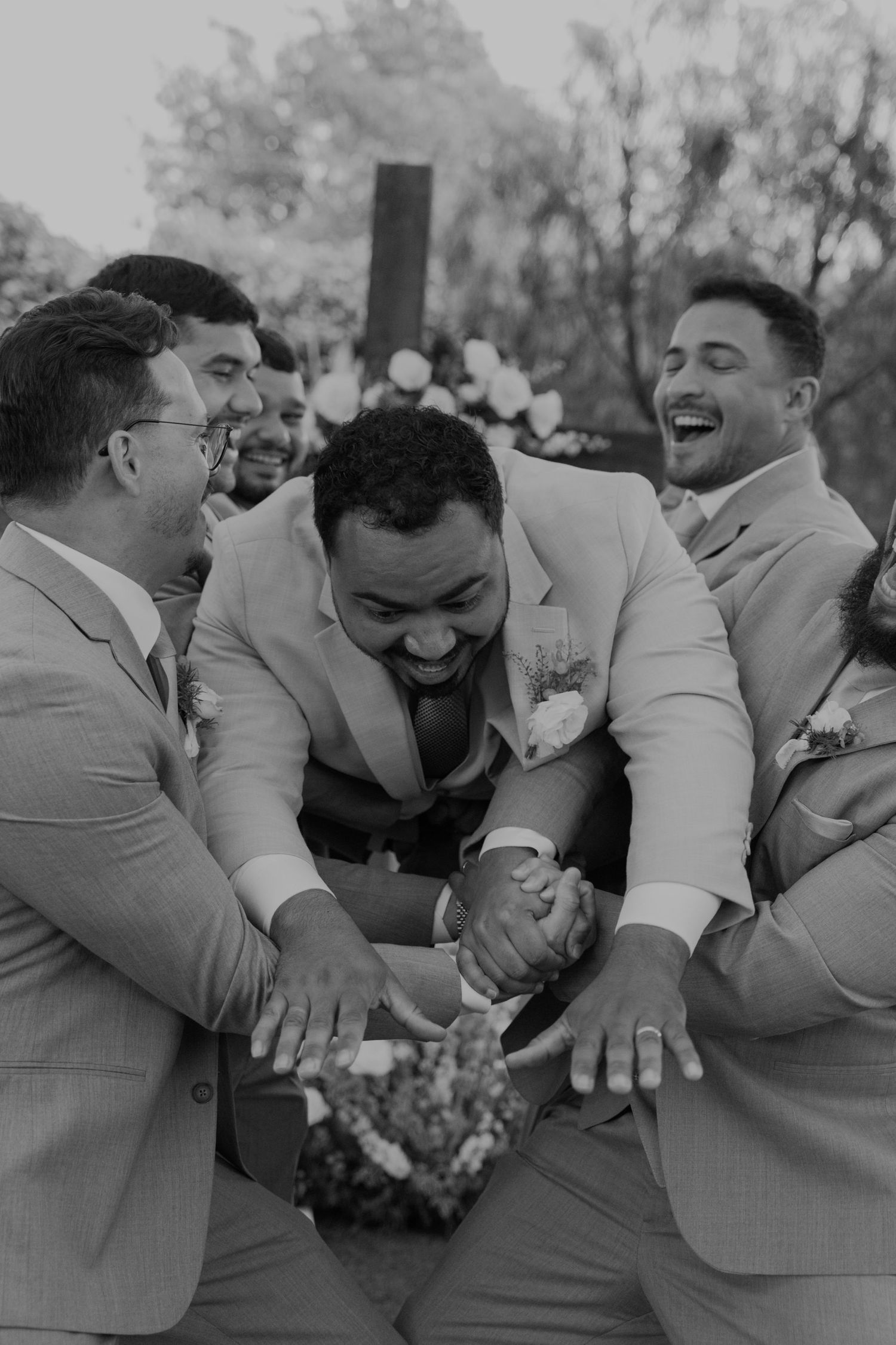 Groomsmen in beige suits laugh joyfully together in black and white candid wedding photo outdoors.