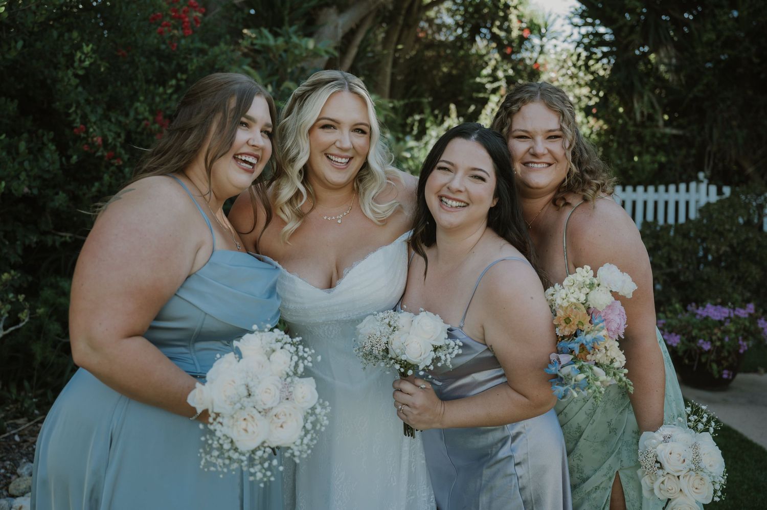Four bridesmaids in blue-gray dresses smile together holding white floral bouquets in a garden setting.