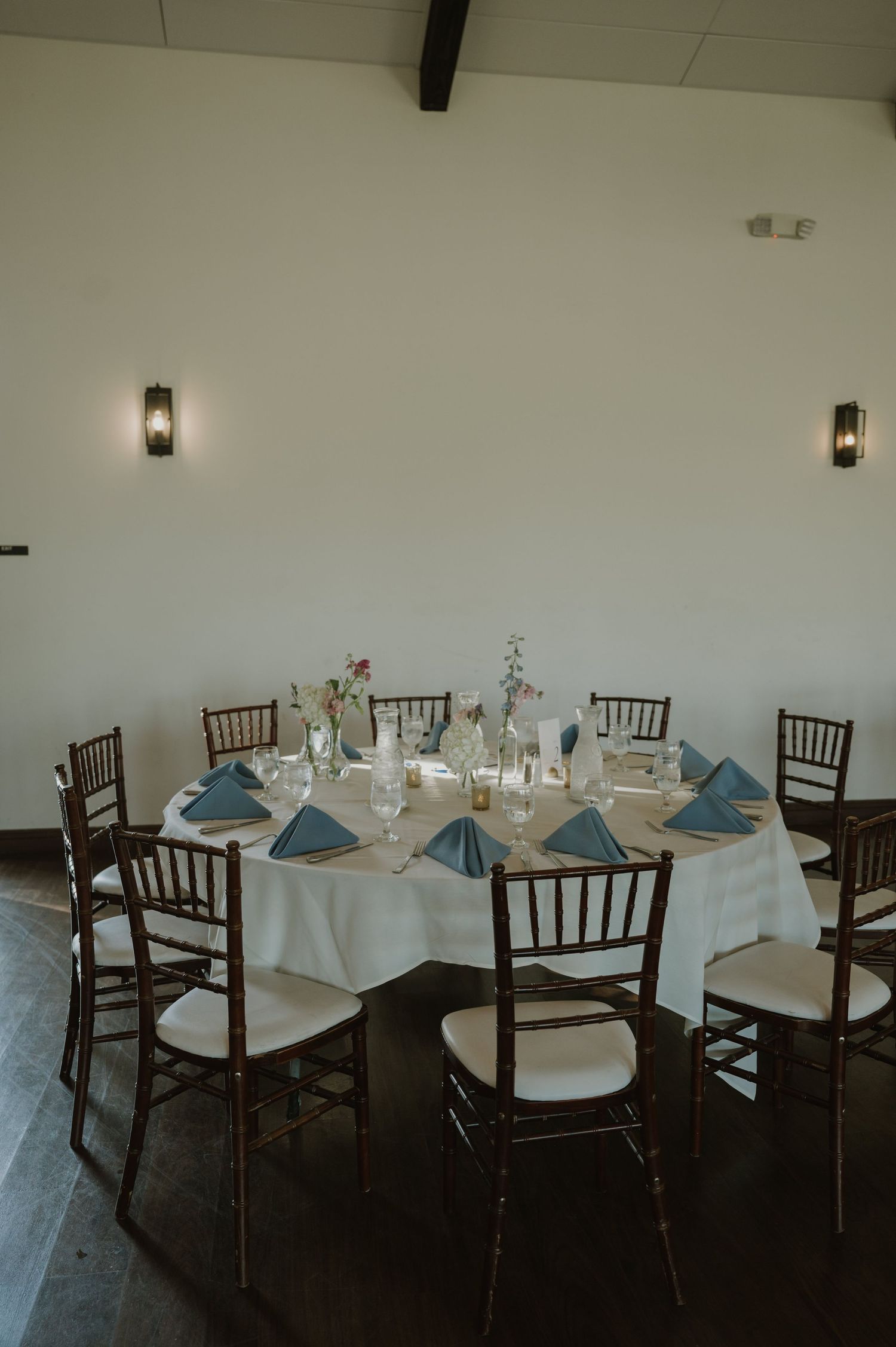 Elegant wedding reception table with blue napkins, floral centerpieces, and chiavari chairs in a bright venue.