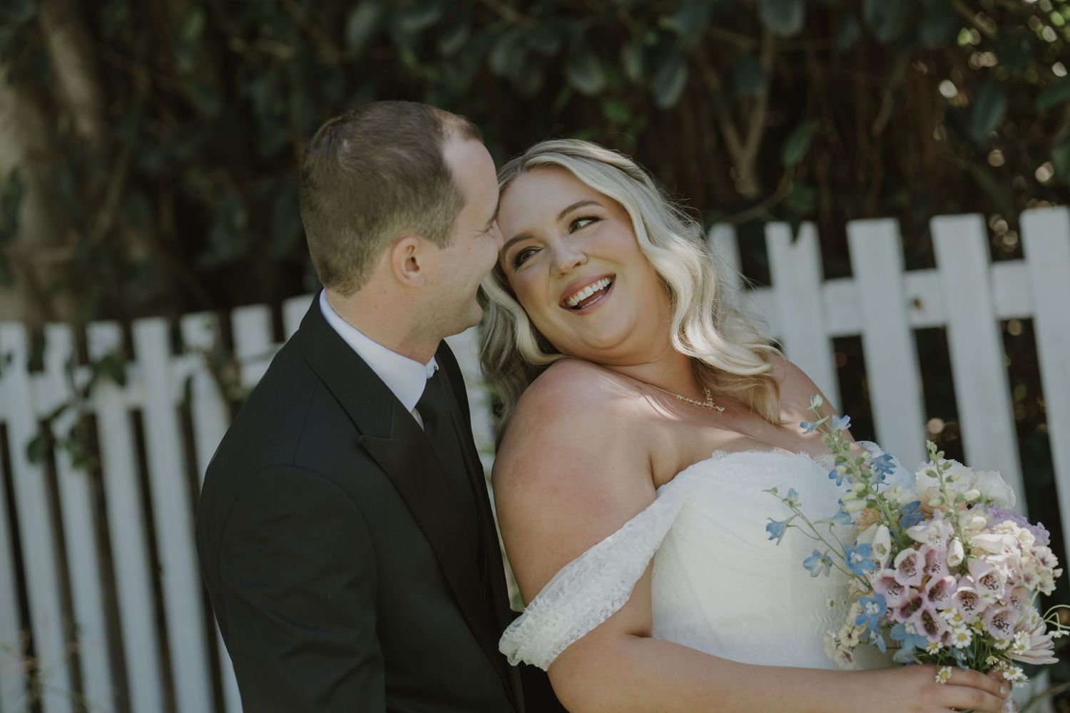 Bride in off-shoulder gown holds floral bouquet as groom in black tuxedo whispers in her ear by white picket fence.