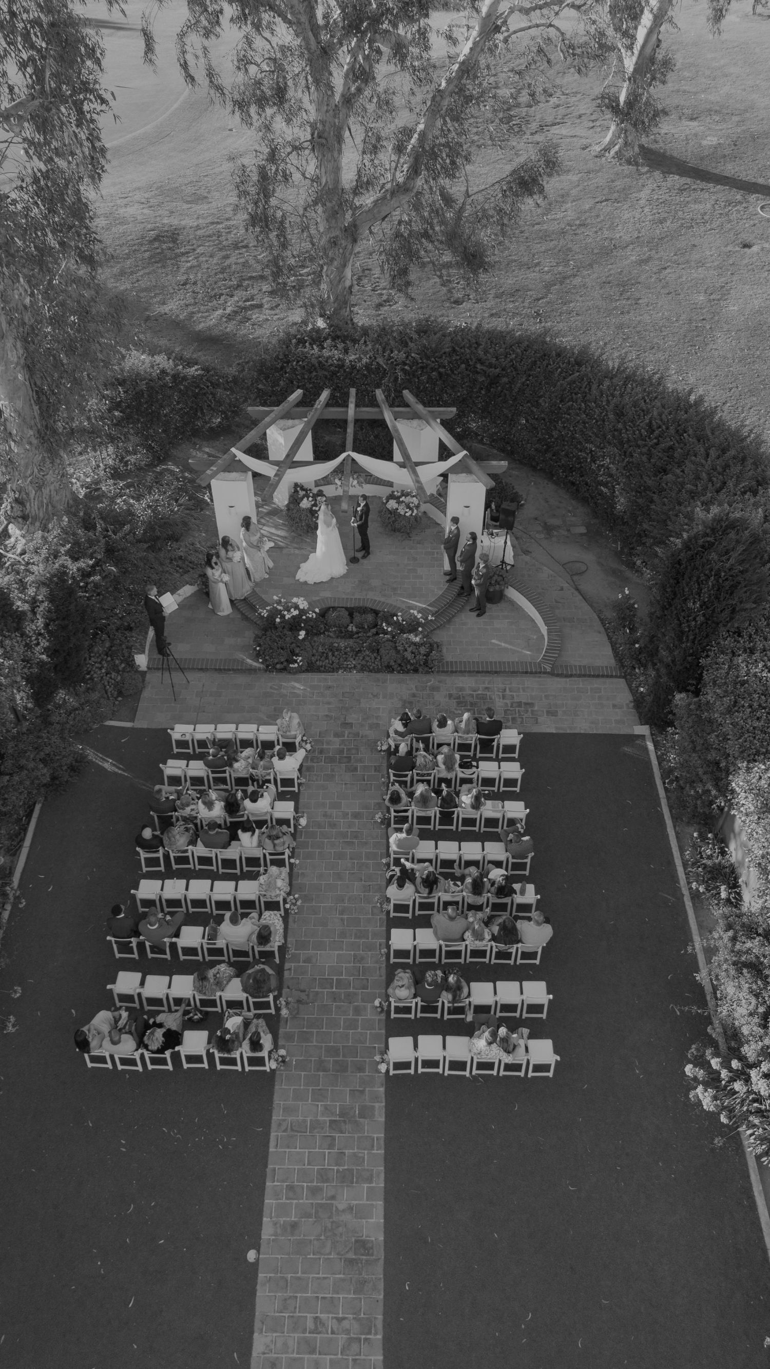 Aerial black and white view of outdoor wedding ceremony with rows of guests seated along brick aisle.