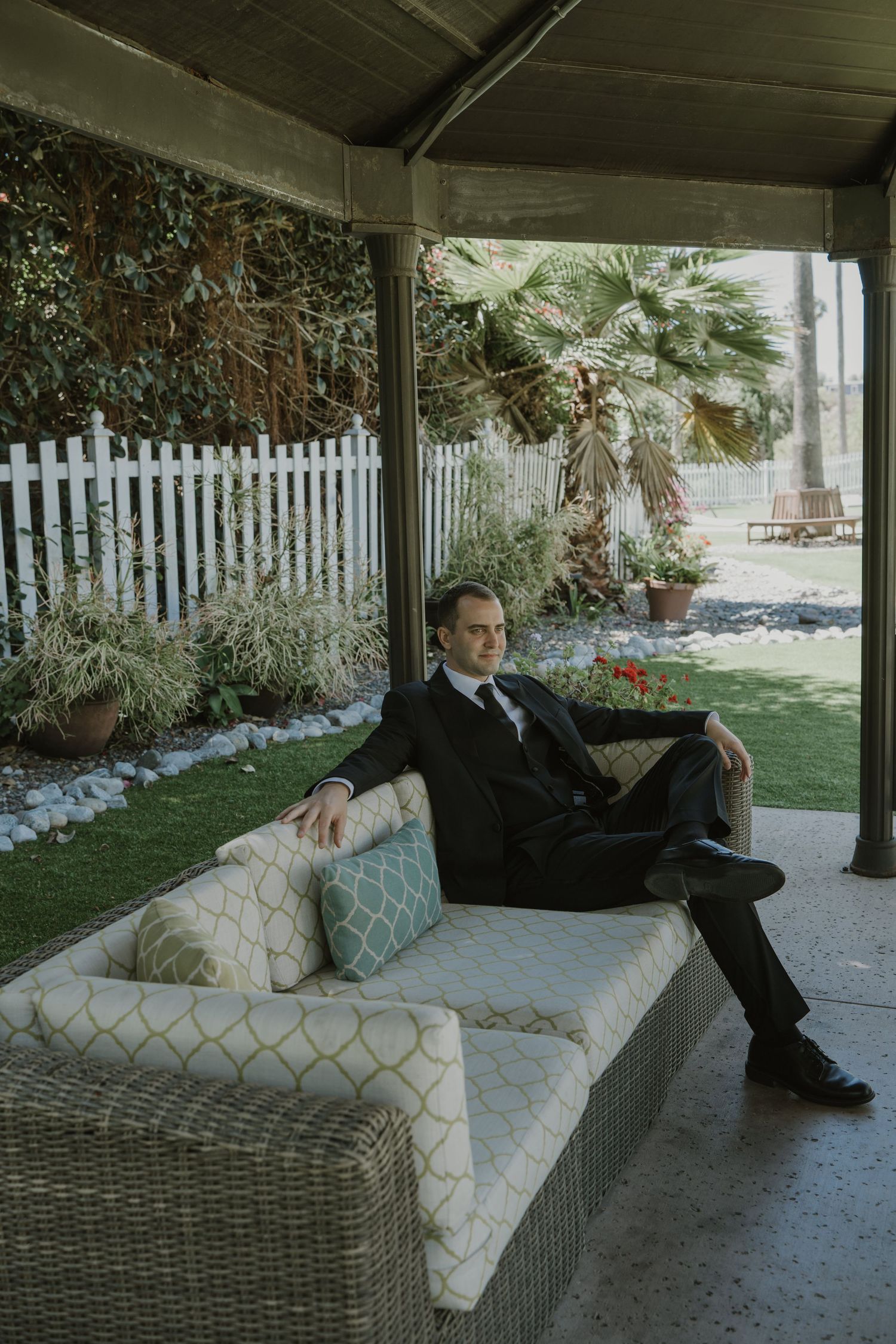 A well-dressed man in a black suit relaxes on a wicker patio sofa surrounded by lush garden greenery.