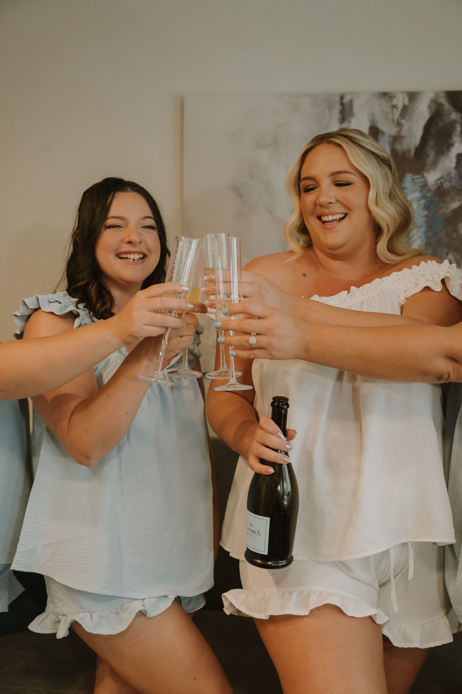 Two women in white pajamas toasting champagne glasses while laughing together.