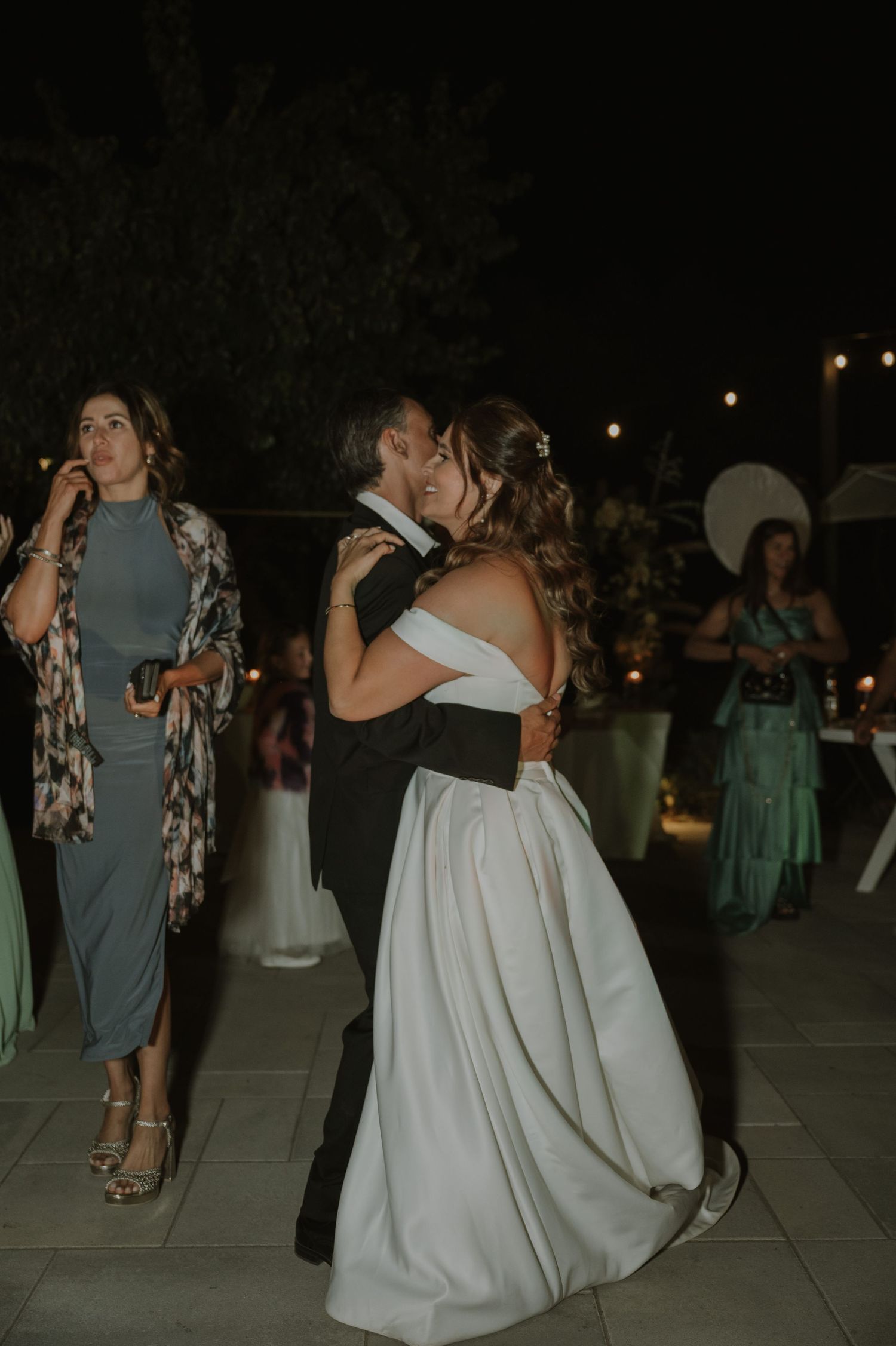 Bride and groom share first dance kiss surrounded by wedding guests at outdoor evening reception.