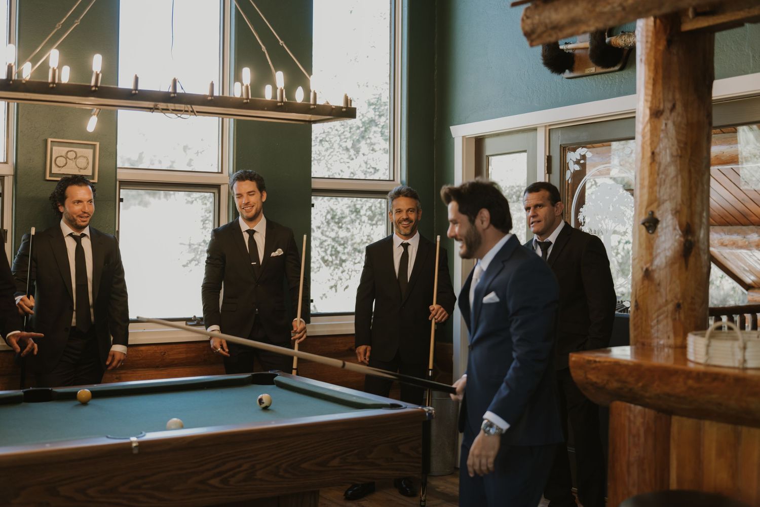 Groomsmen in black suits playing pool in a rustic lodge before a wedding ceremony.