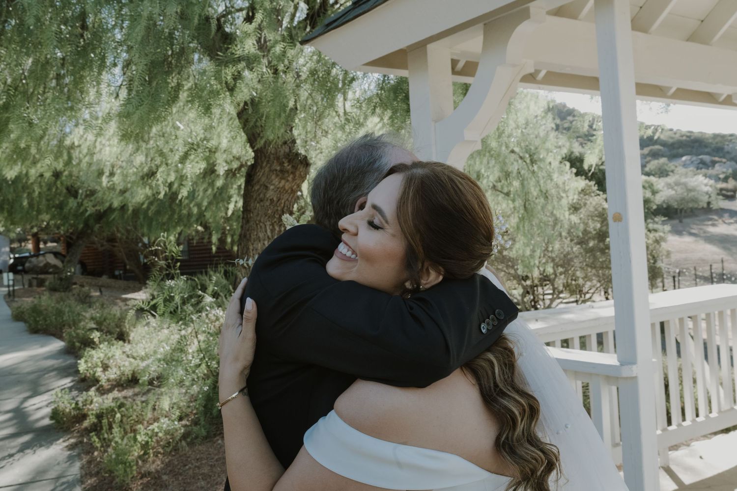 Bride in white gown shares emotional hug with guest under pergola surrounded by trees.