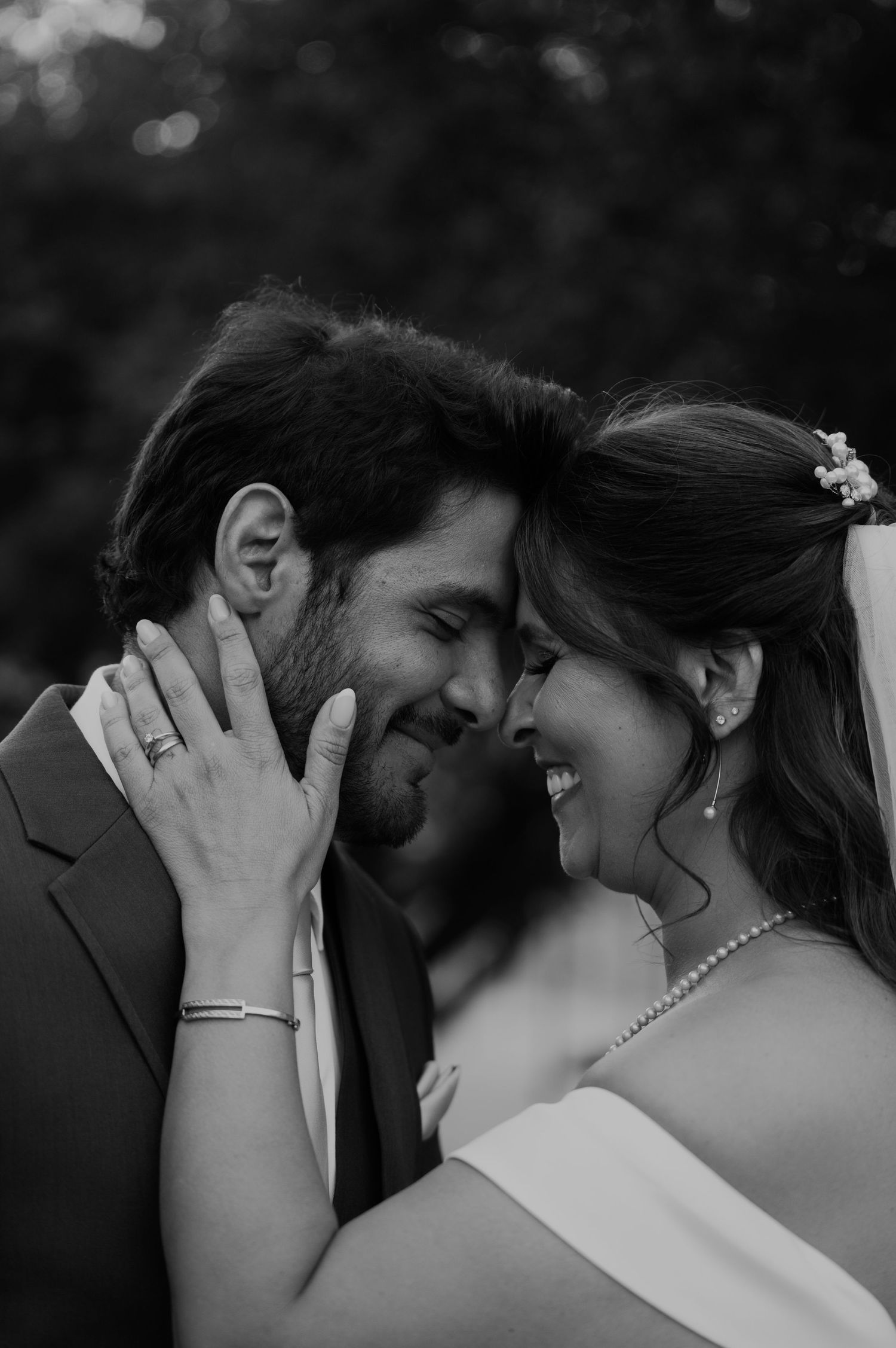 Black and white wedding photo of smiling bride and groom touching foreheads, bride wearing pearl necklace and ring.