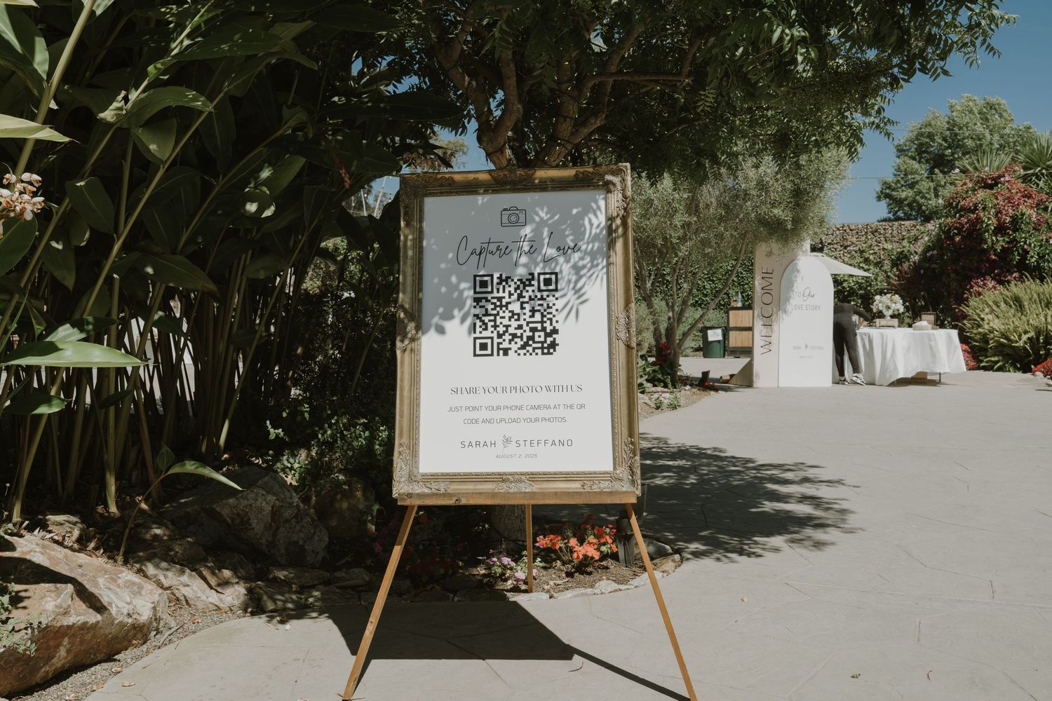 A wooden easel displaying a QR code wedding sign surrounded by lush greenery at an outdoor venue.