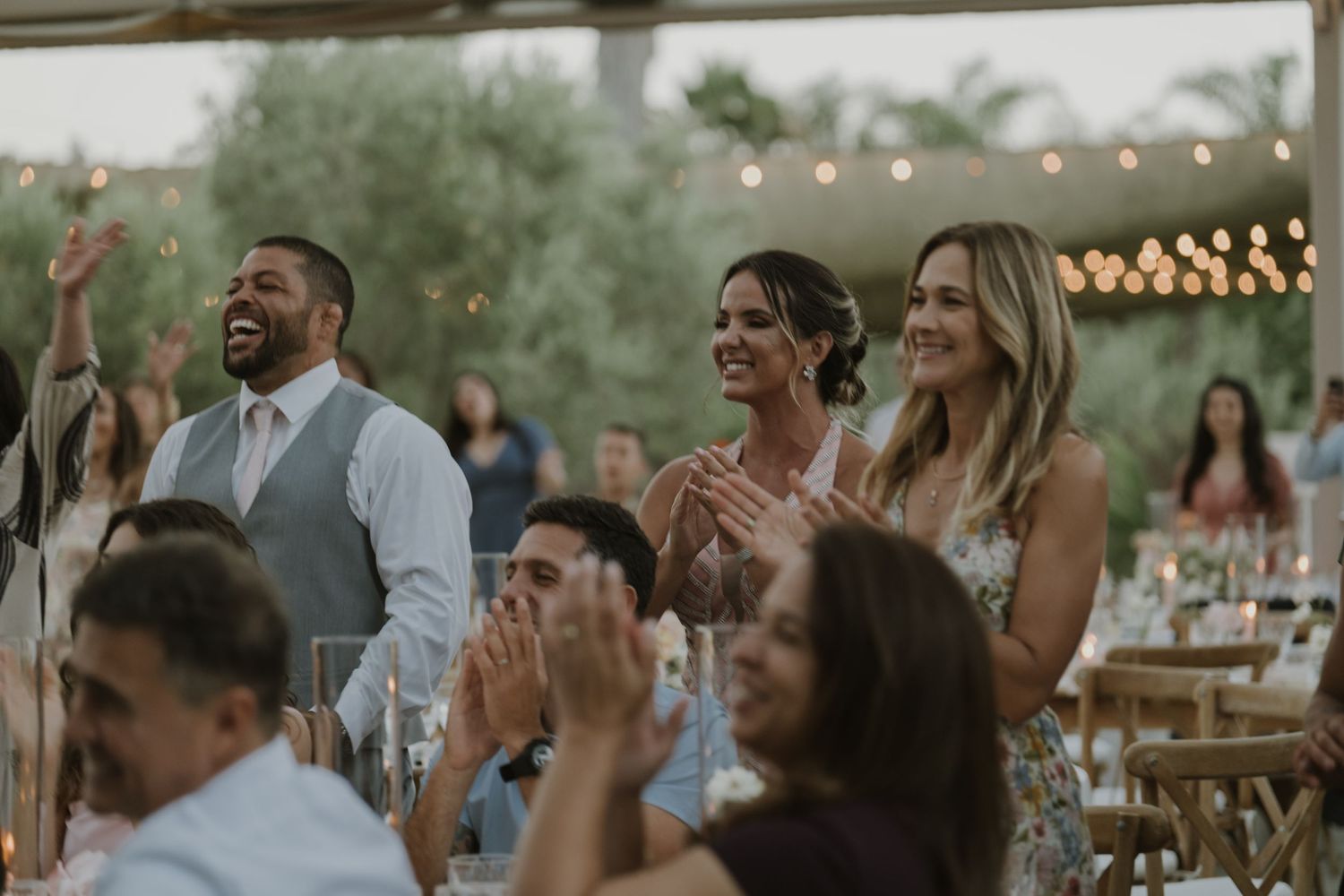 Guests clapping and laughing at an outdoor wedding reception under string lights.