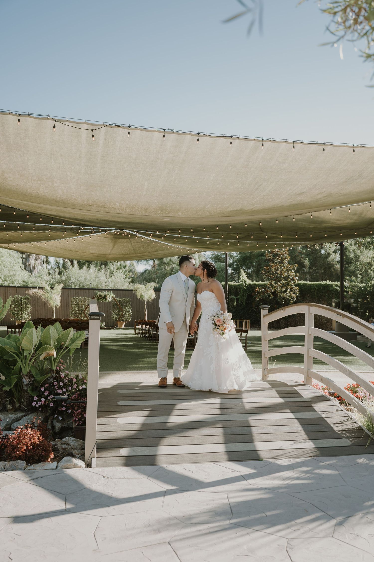 Newlyweds sharing a kiss on a wooden bridge under a canopy with string lights at an outdoor wedding venue.
