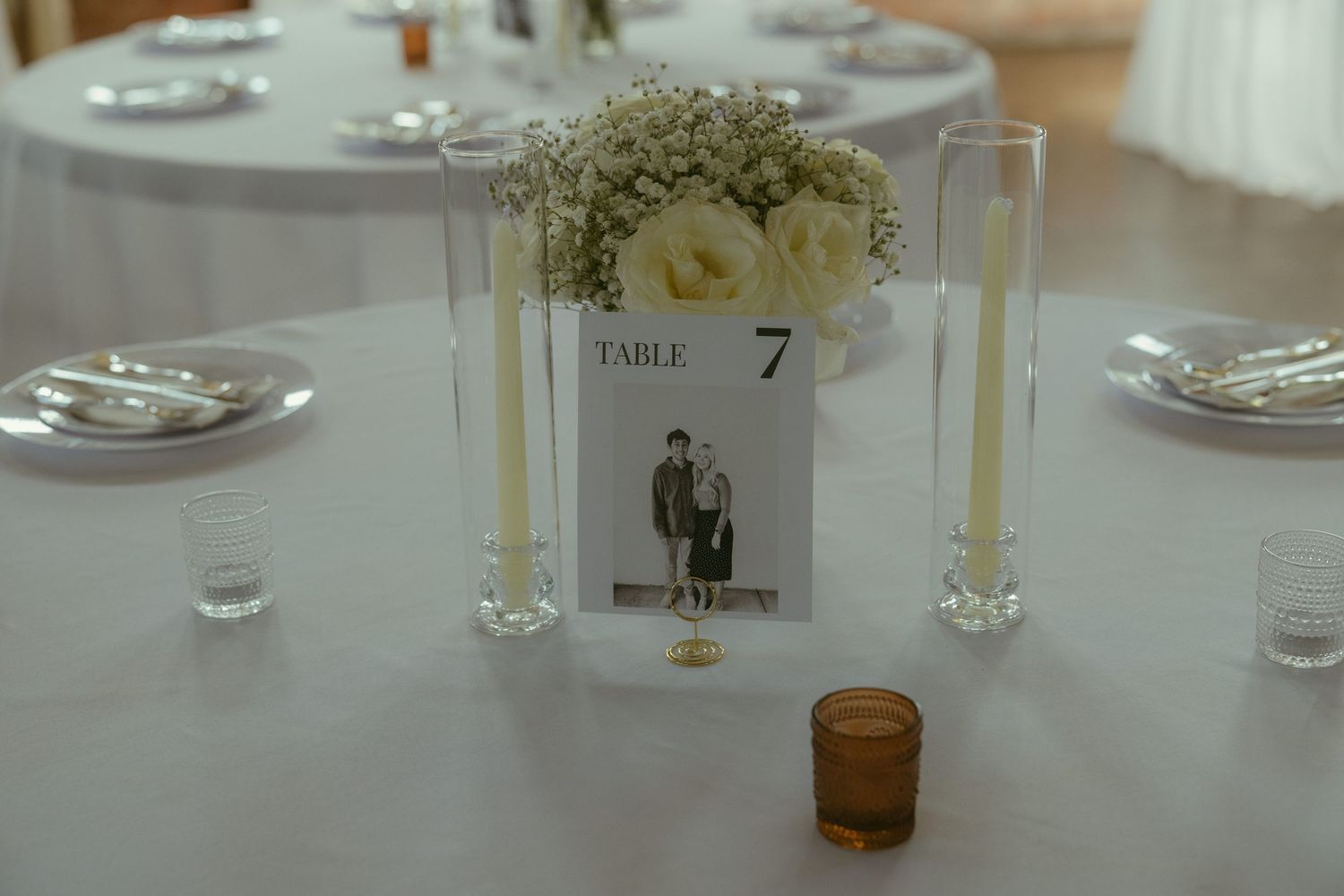 Elegant wedding reception table 7 with white floral centerpiece, candles, and a couple's photo card on white linen.