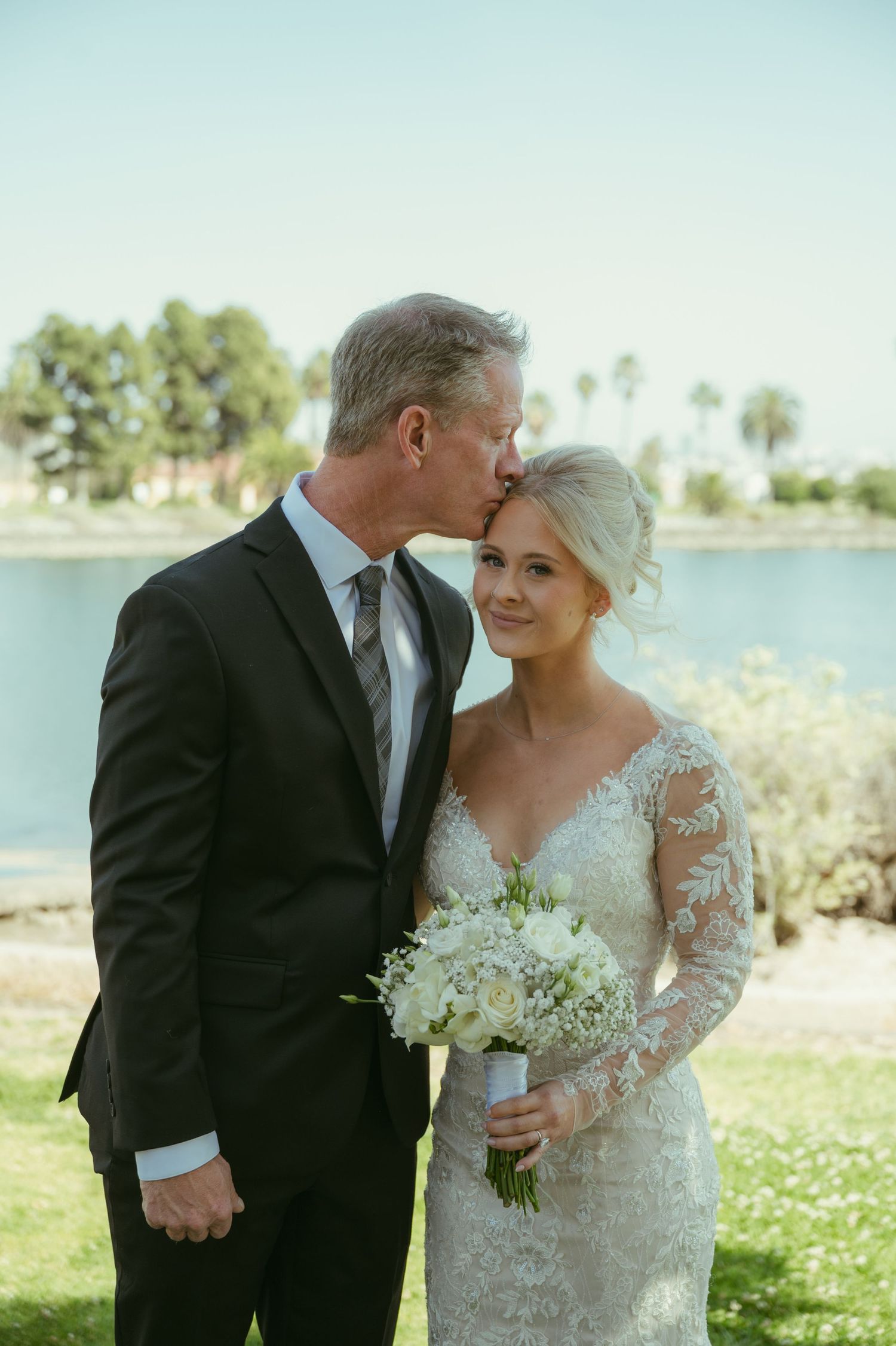 Father kissing bride on forehead as she smiles holding white bouquet, standing near scenic lake with palms.
