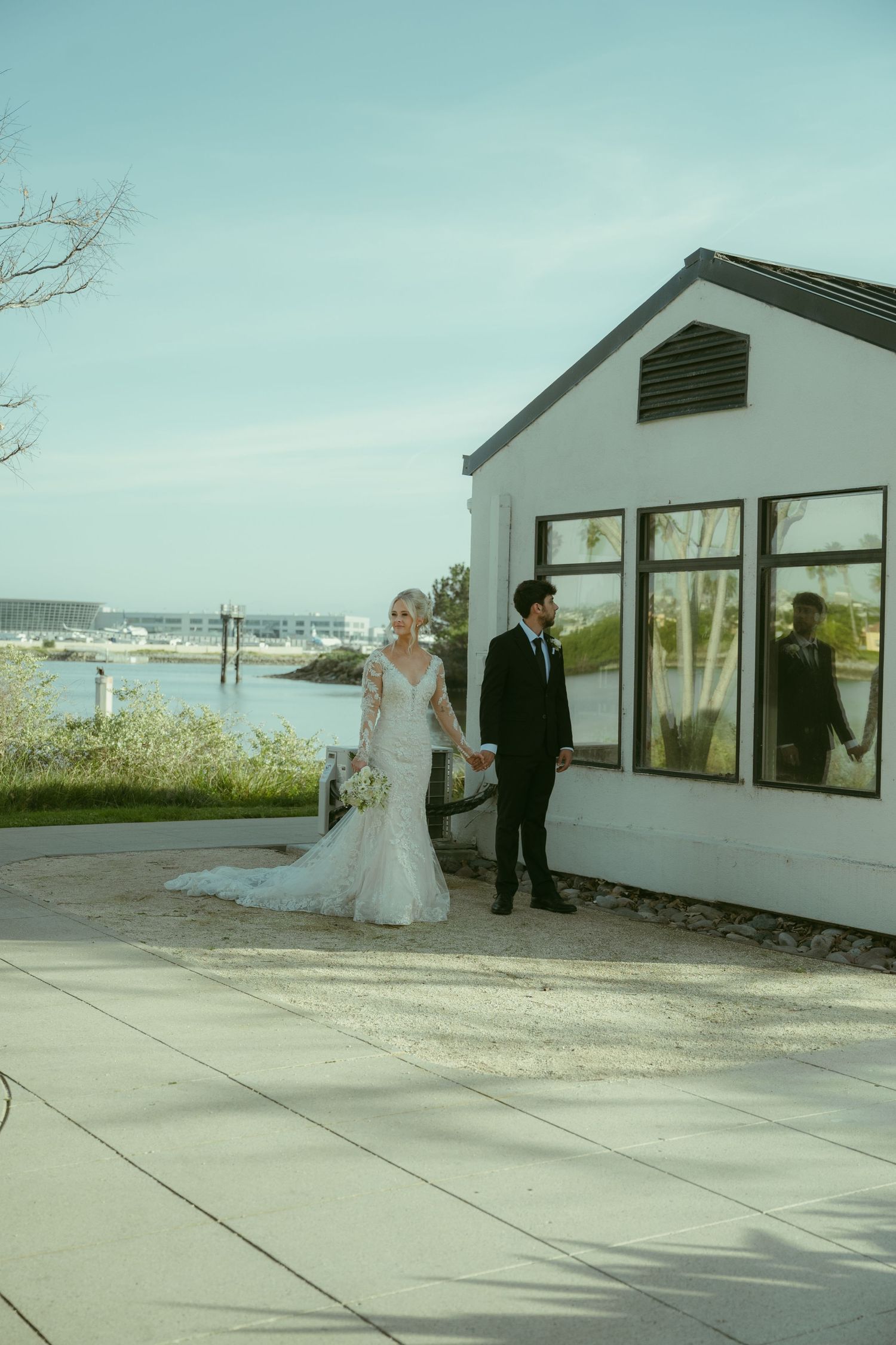 Bride and groom walk hand in hand near a waterfront venue on their wedding day.