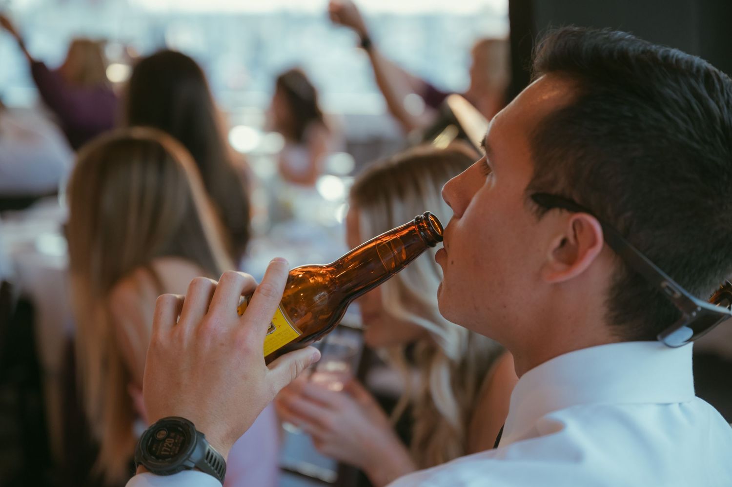 Wedding guest in white shirt drinks from beer bottle at outdoor reception celebration.