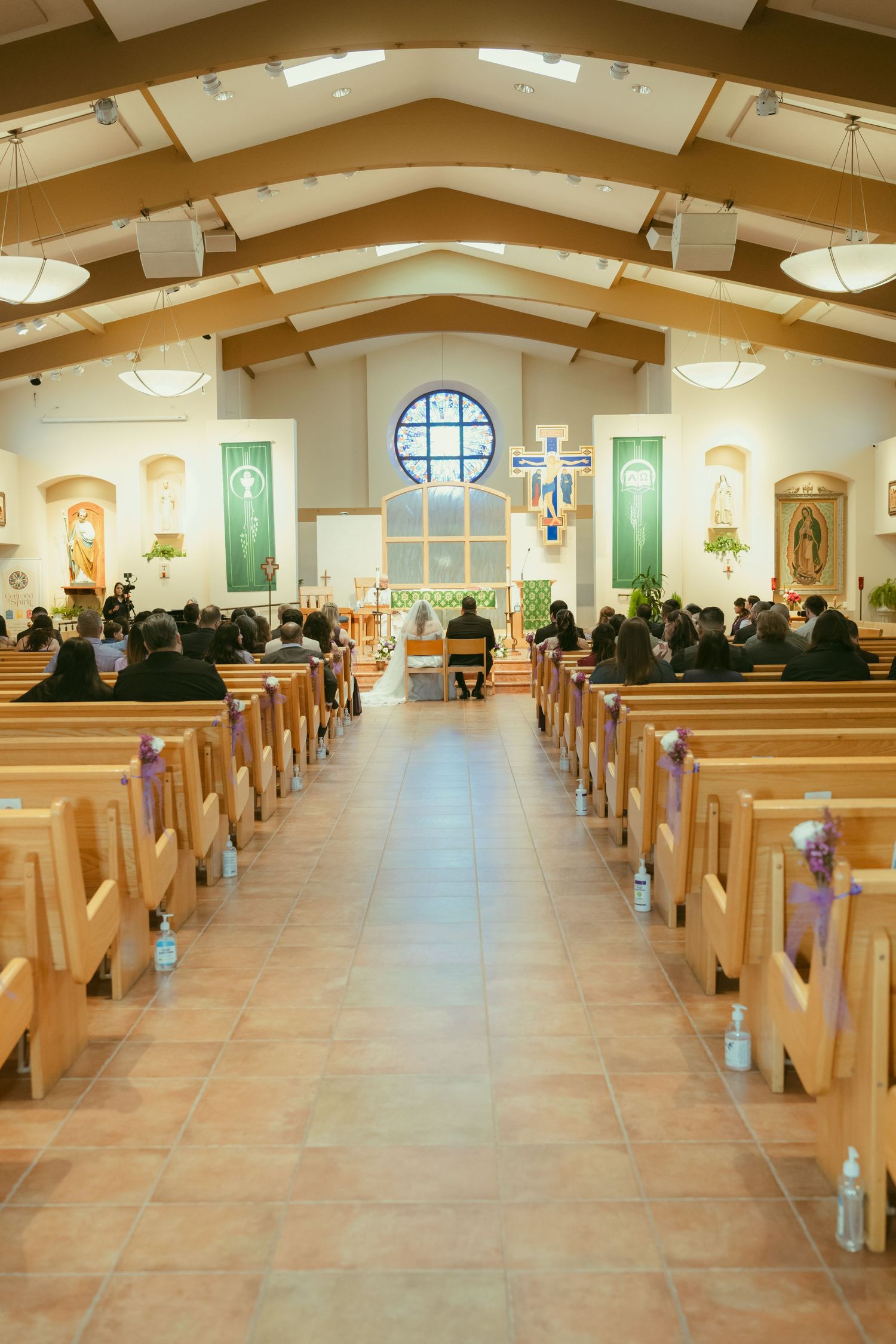 Church interior with wooden pews filled with guests during a wedding ceremony, featuring a cross and stained glass window.