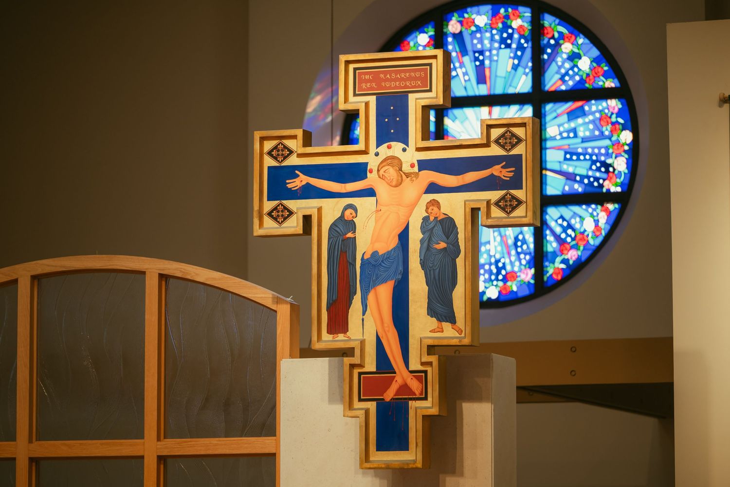 Colorful San Damiano crucifix icon displayed in a church with a vibrant stained glass rose window backdrop.