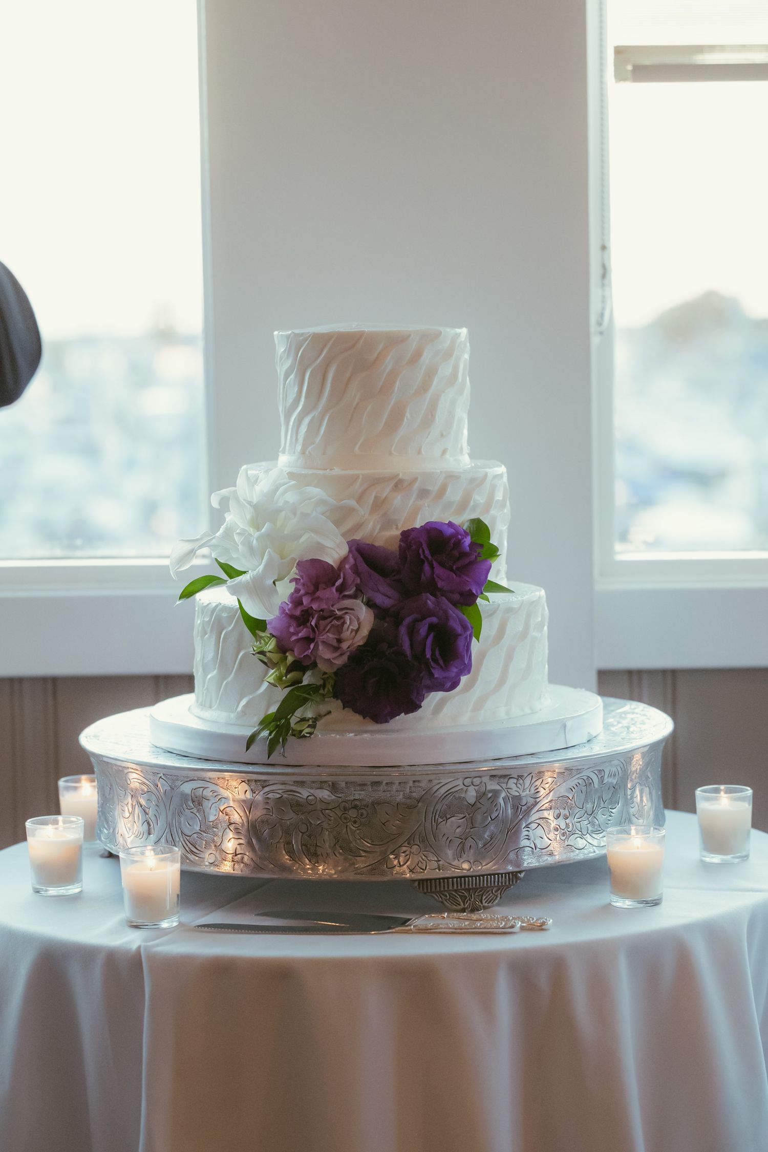 Three-tier white wedding cake with purple flowers on silver stand surrounded by candles.