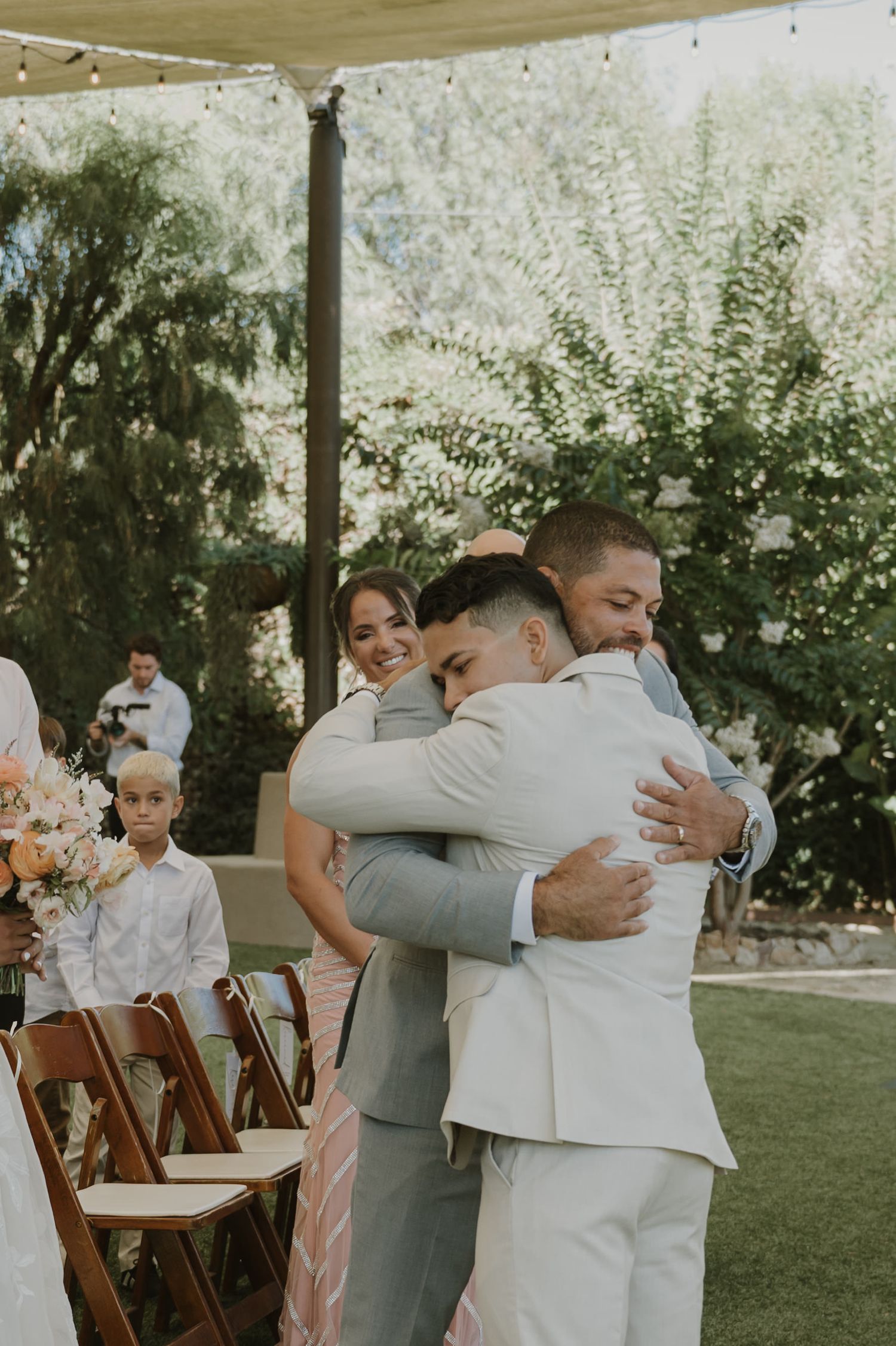 Two grooms share an emotional hug during an outdoor wedding ceremony surrounded by guests and lush greenery.