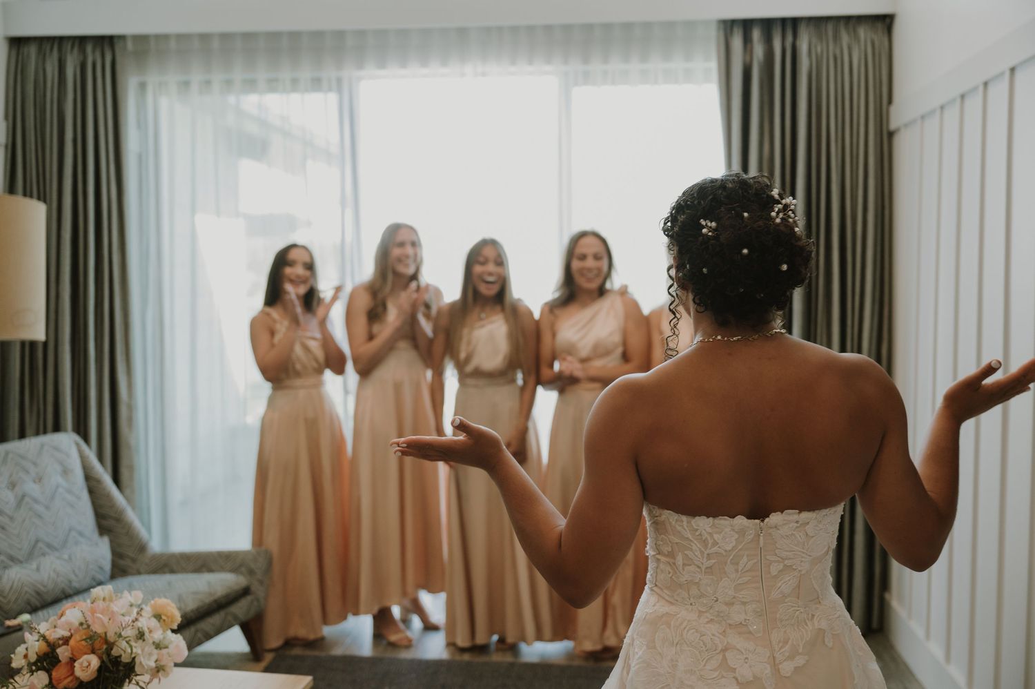 Bride in lace gown reveals her look to four cheering bridesmaids in champagne dresses.