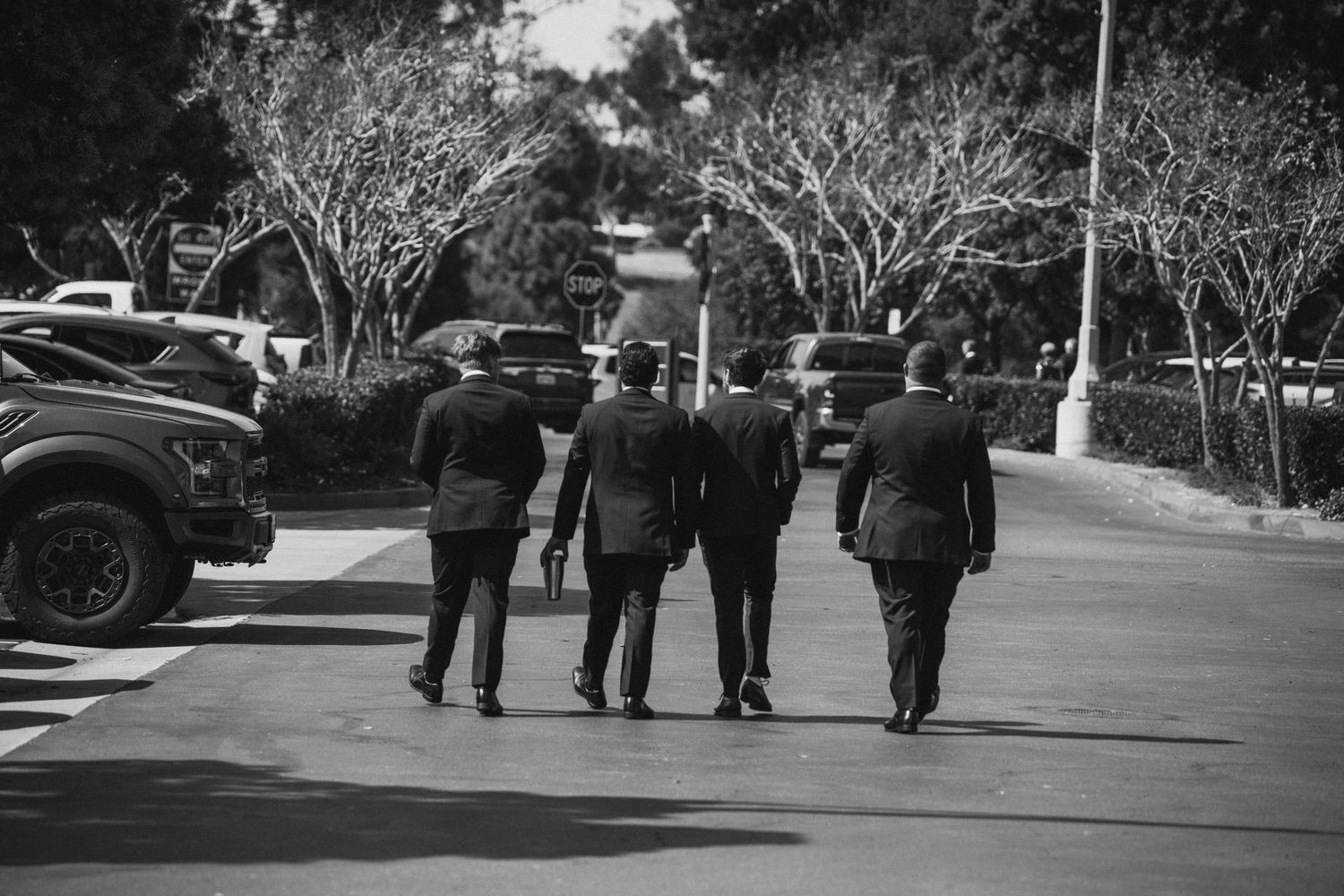 Four men in formal suits walking together down a tree-lined path in a black and white photo.